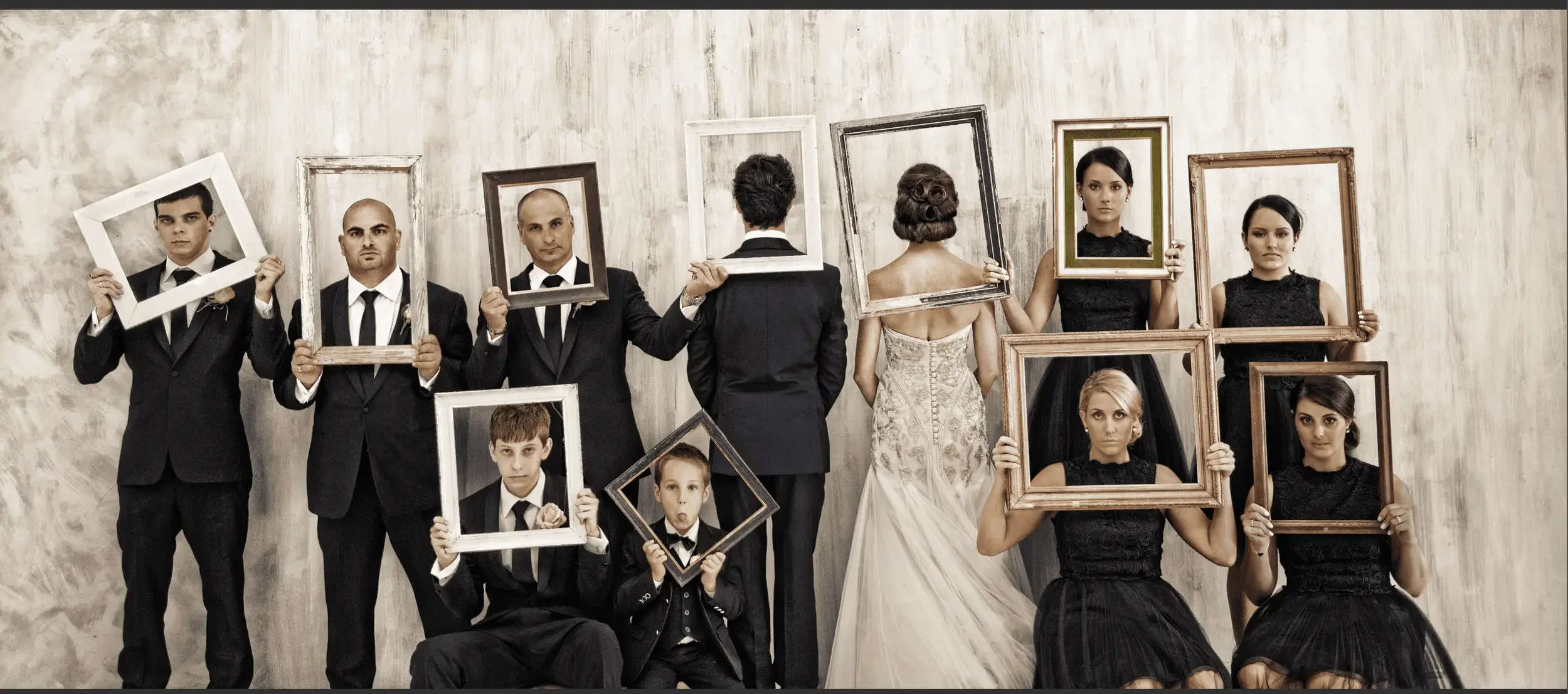 A group of people in formal attire holding empty picture frames while facing a textured light-colored wall.