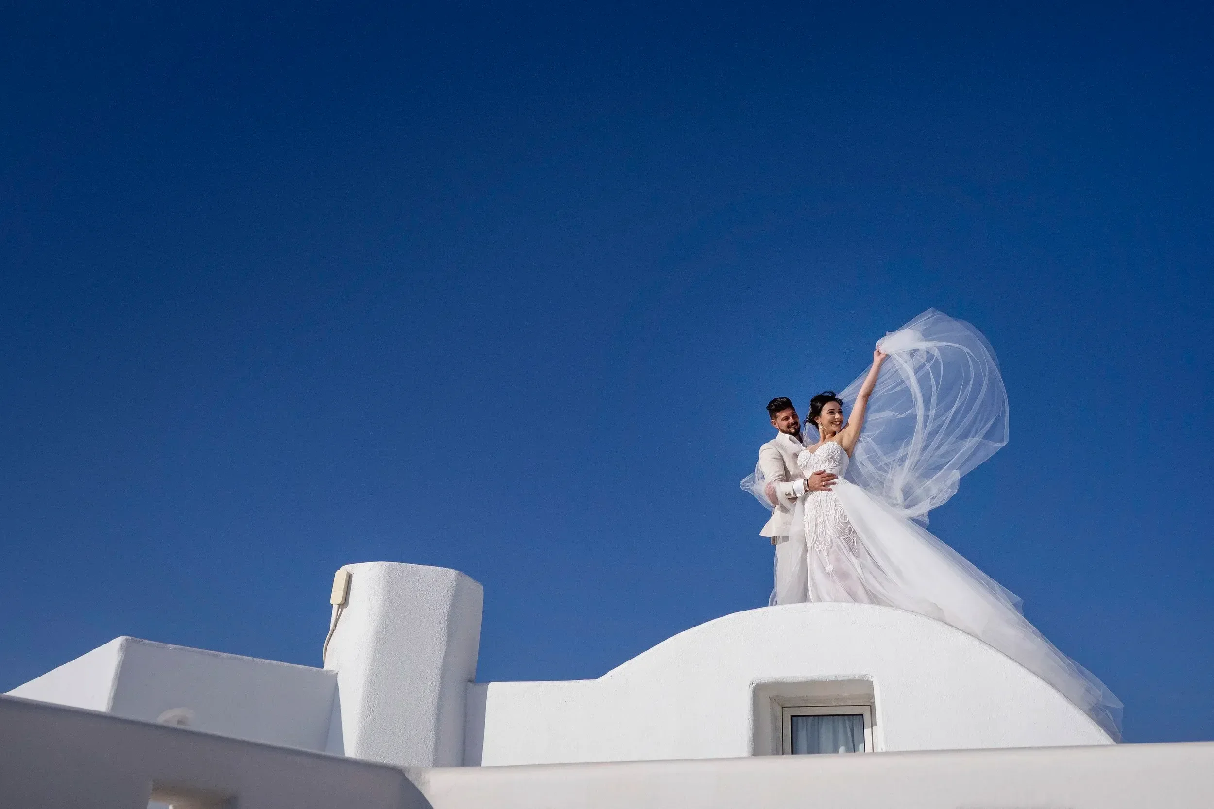 A bride and groom in wedding attire standing on a white rooftop with a deep blue sky in the background, celebrating their wedding moment.