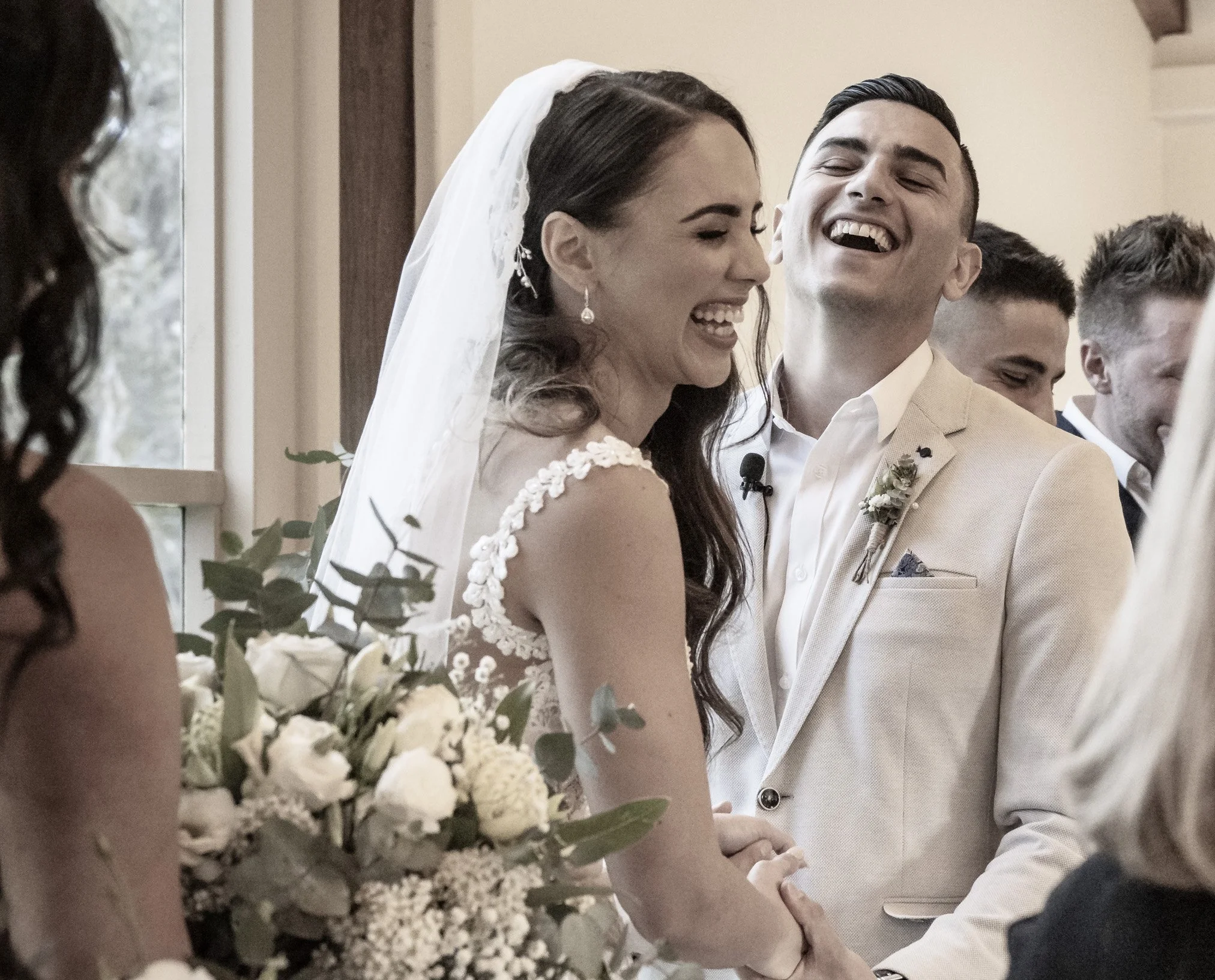 A bride and groom laugh during their wedding ceremony, holding hands, with bridesmaids and groomsmen in the background.