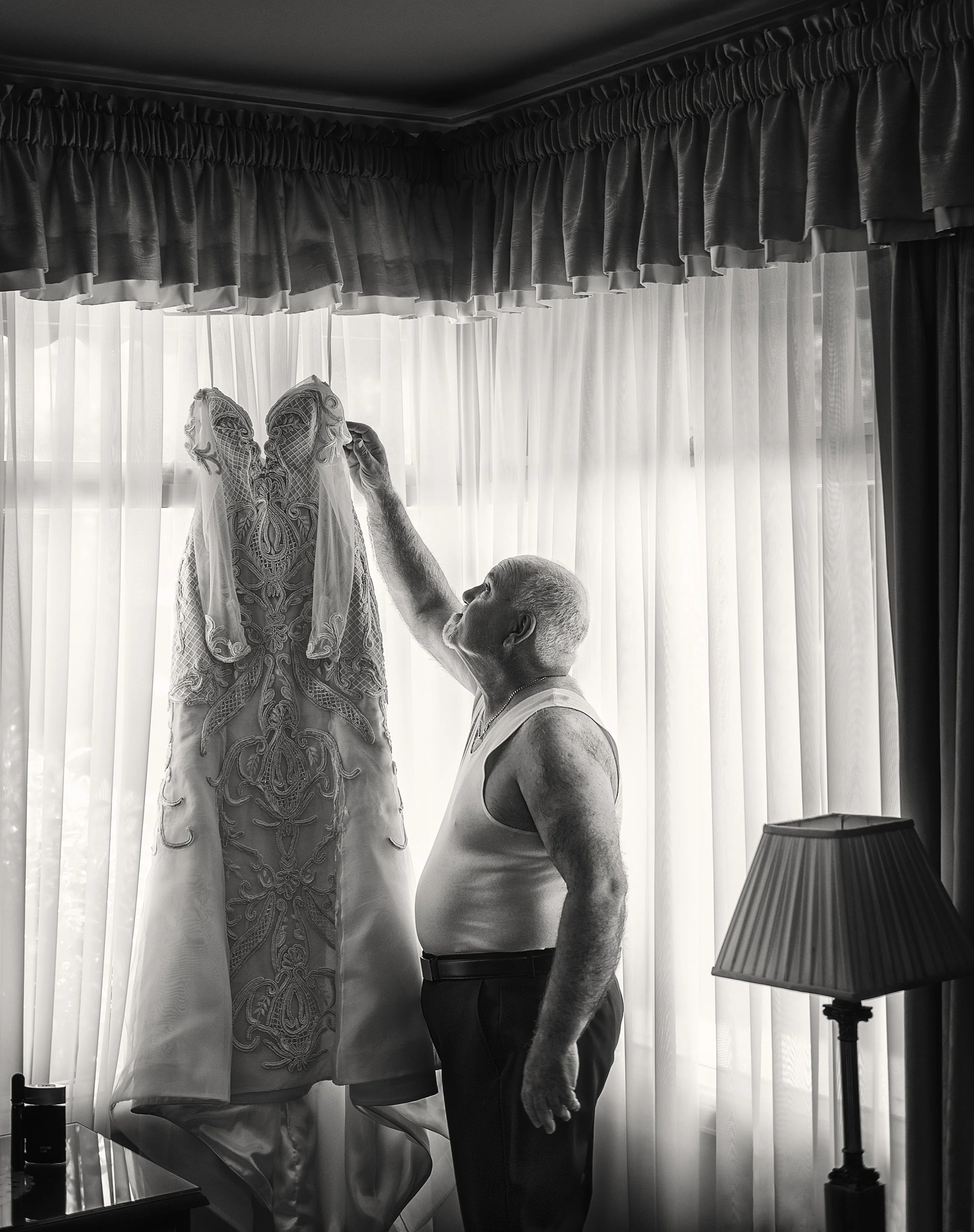 An elderly man in a tank top and dark pants looks at a wedding dress hanging near a window with curtains, touching the dress with his right hand.