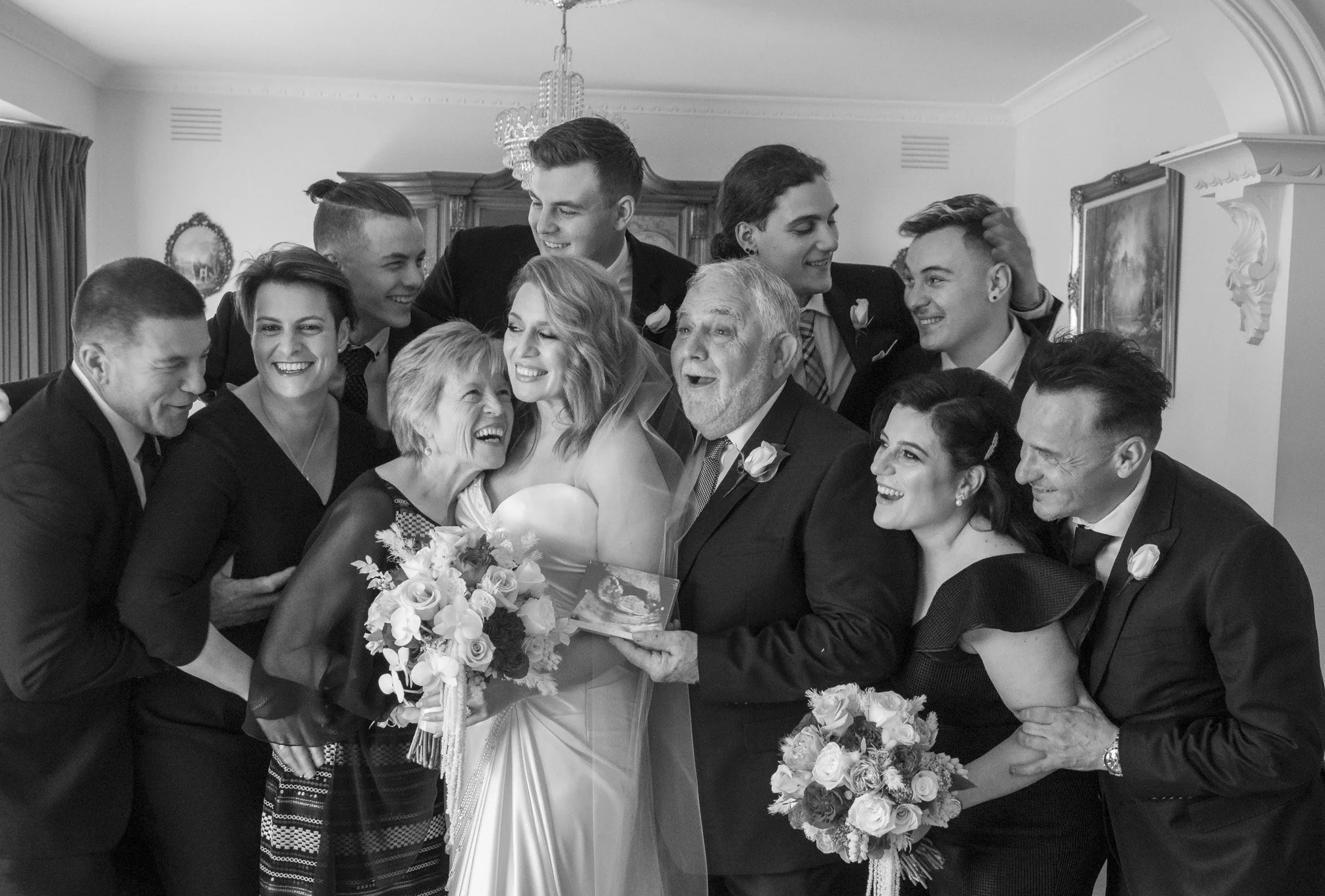 A group of people at a wedding, gathered closely and smiling, holding bouquets, with a bride and groom in the center.