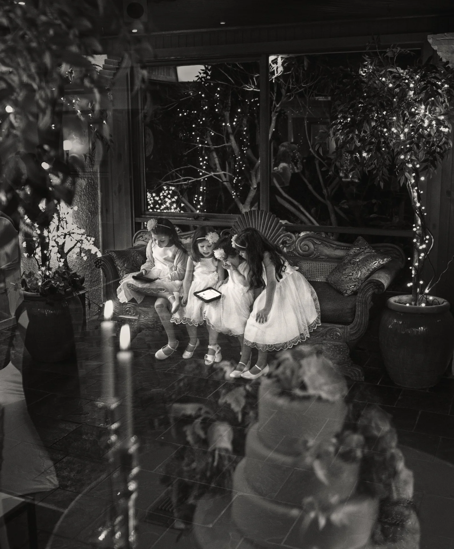 Four young girls in white dresses sitting on a sofa, looking at tablets, with decorative flowers in their hair, in a cozy, decorated indoor setting with lights outside through the window.