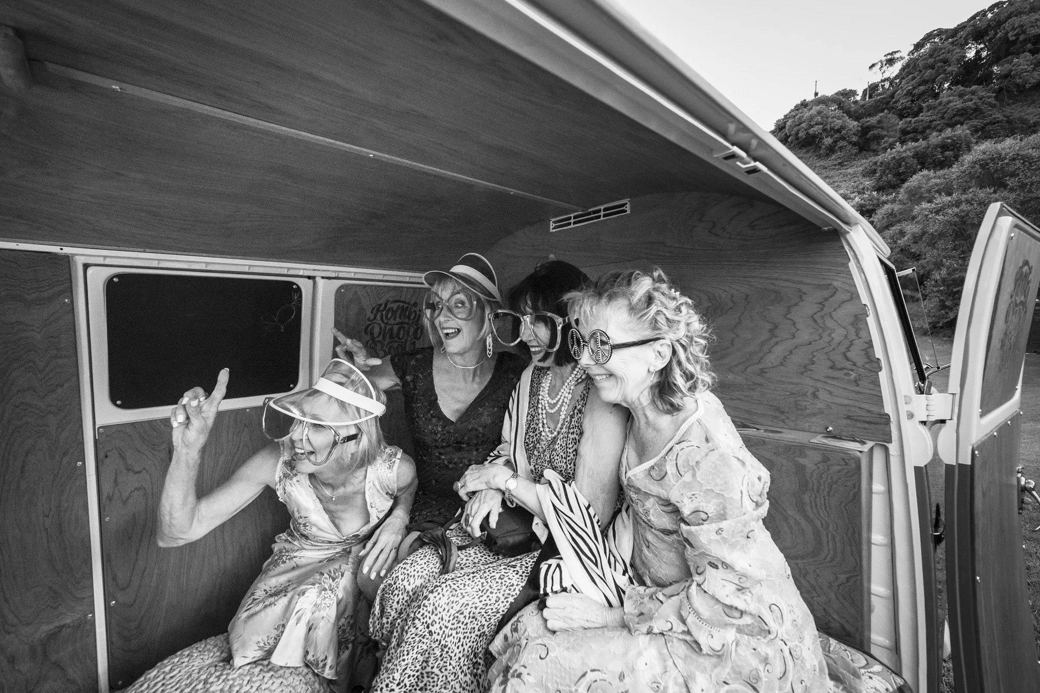 Five women wearing large novelty glasses and hats, smiling and laughing inside a vintage van with wooden interior, outside on a hillside.