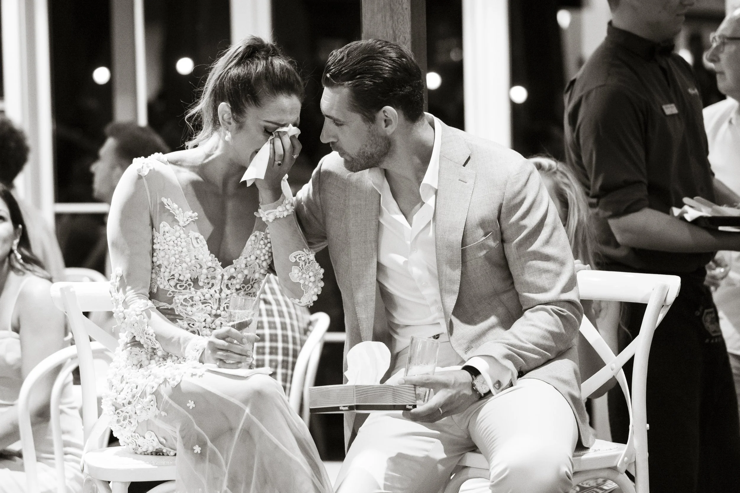 A man proposing to a woman at a wedding reception, she is crying and holding a glass of champagne, while he holds a ring box and touches her face.