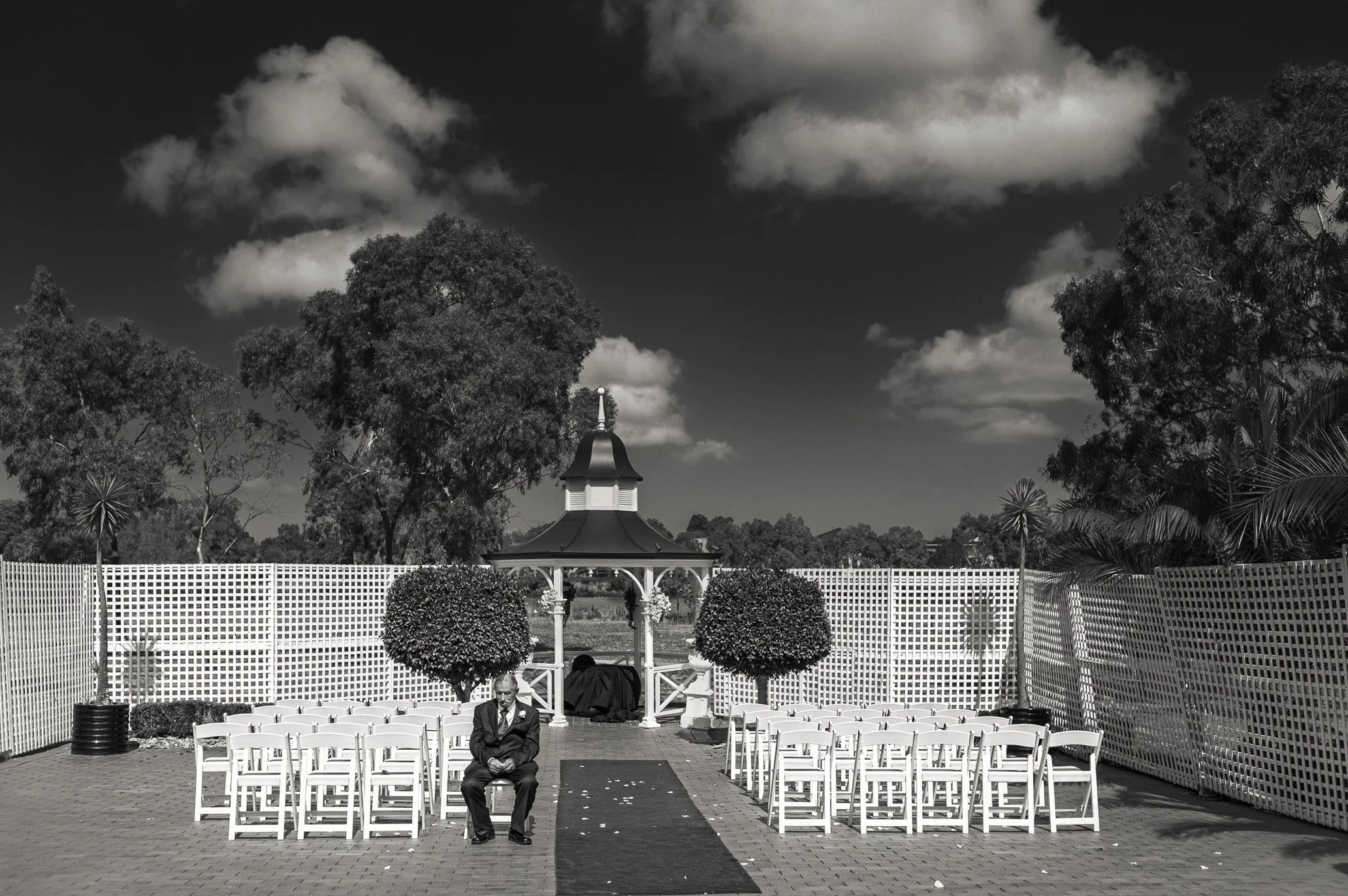 An outdoor wedding ceremony setup with white chairs arranged in rows on either side of an aisle. There is a gazebo in the background surrounded by manicured bushes and trees, with a man sitting in the front row. The sky is partly cloudy.