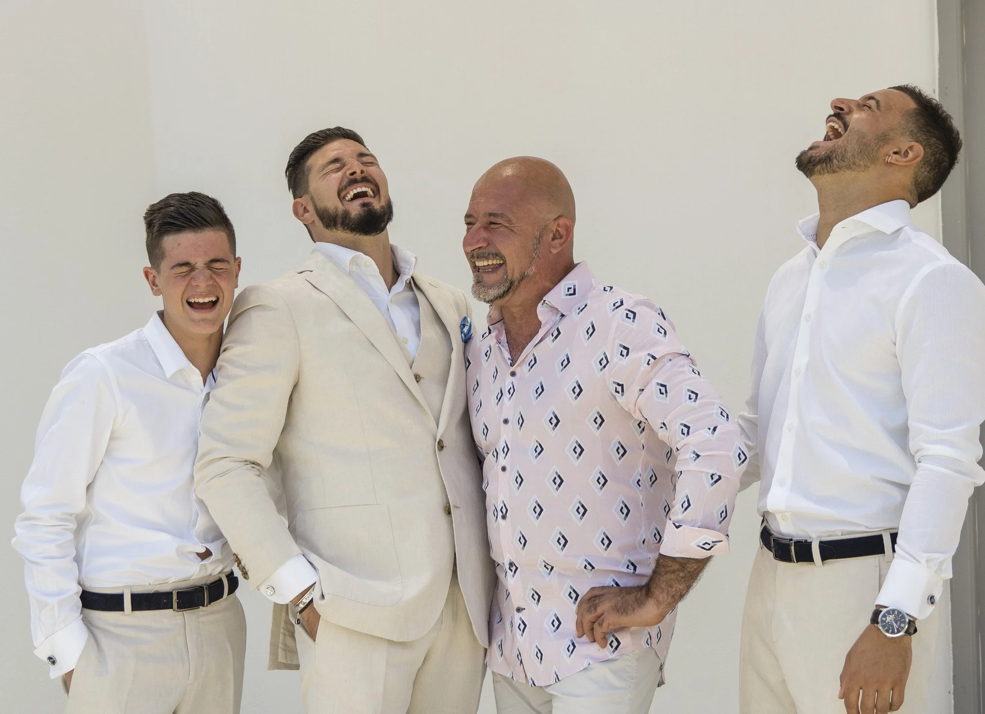 Five men laughing and enjoying each other's company, dressed in formal and semi-formal attire, against a plain white background.