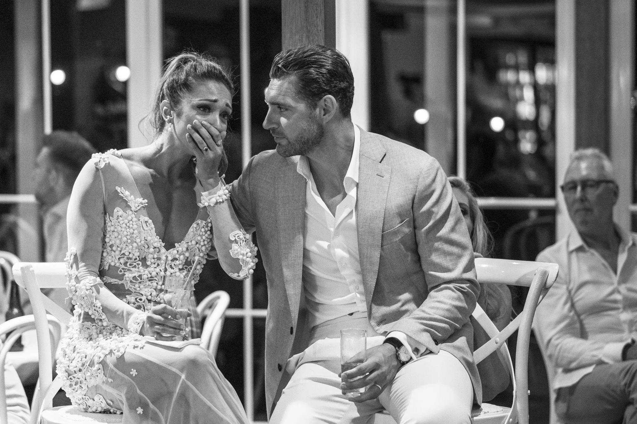 A woman in an embroidered dress sitting next to a man in a blazer, both holding drinks, with the woman appearing emotional and the man looking at her with concern, in a social setting with other guests in the background.