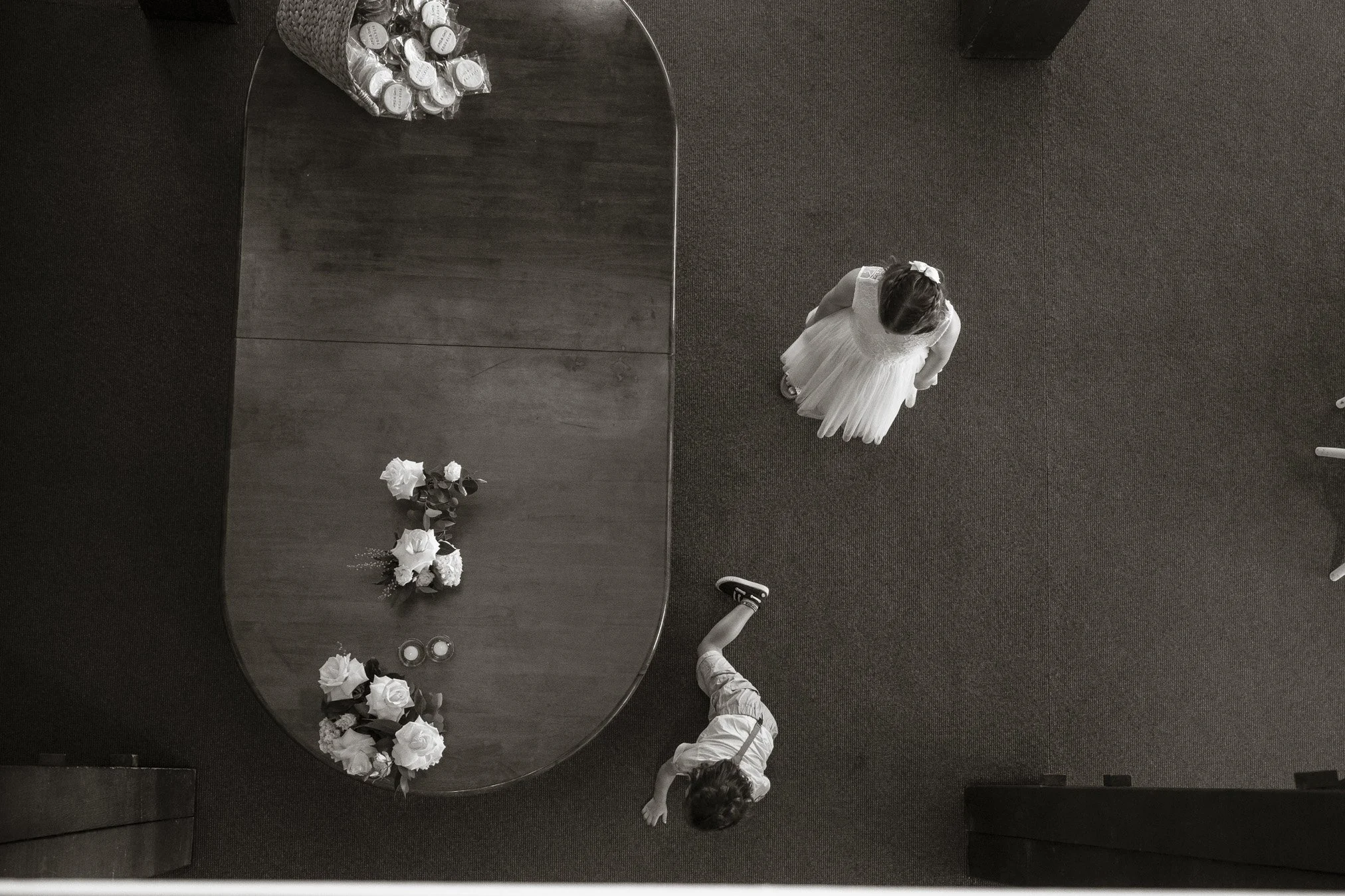 A girl in a white dress, possibly a flower girl, standing on a carpeted floor, viewed from above. A young boy in light-colored clothes is approaching her from behind, and a table with flower arrangements, candles, and small wrapped boxes is nearby.
