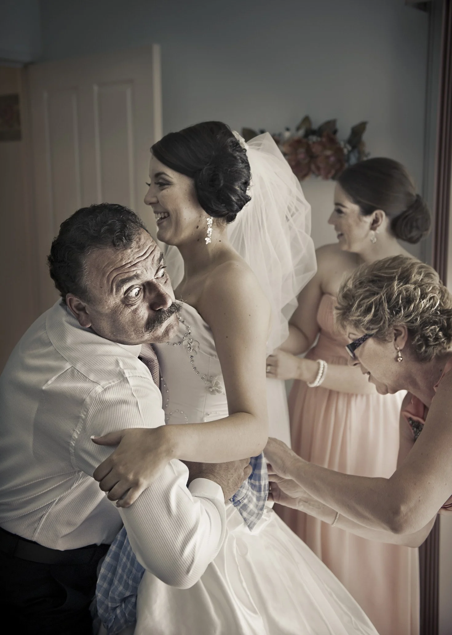 A bride in a wedding dress smiling and hugging a man, likely her father, who is holding her and looking up at her. Two women, possibly bridesmaids or family members, stand in the background, smiling and assisting.