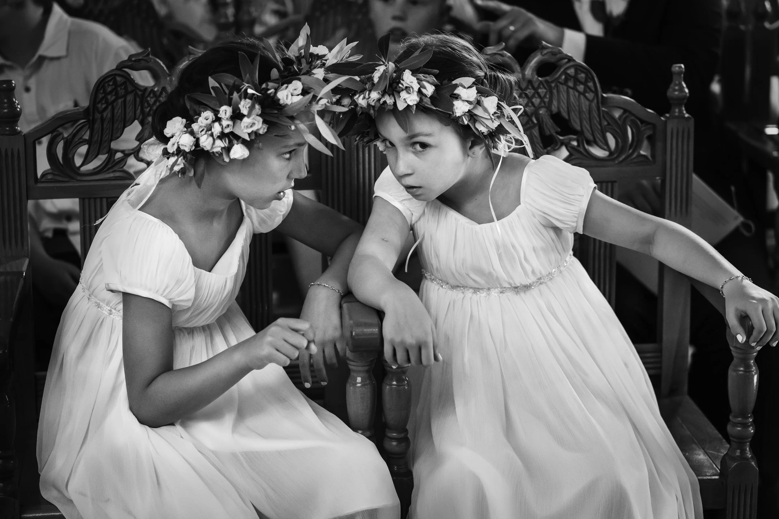 Two young girls wearing white dresses and floral crowns sitting and leaning towards each other in a church or formal setting.