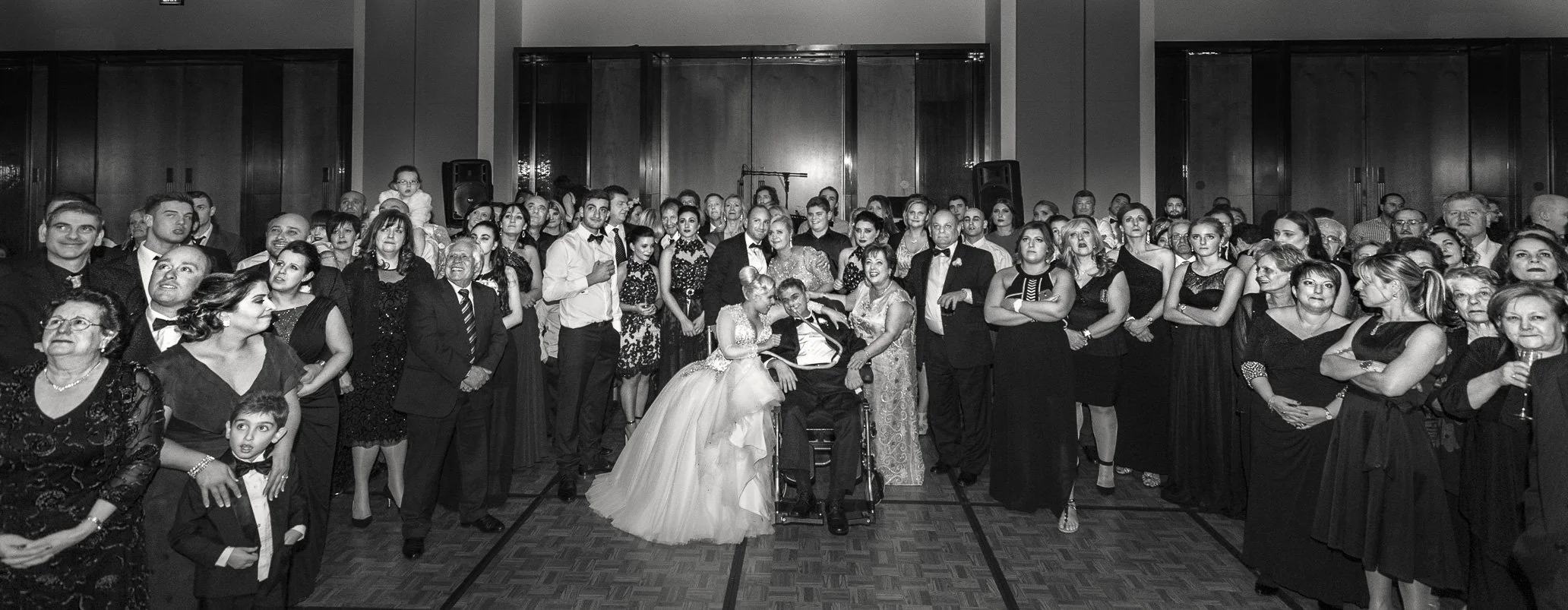 A large group of people dressed in formal attire gathers in a ballroom for a celebration or event. The group includes men, women, children, and elderly individuals, with some smiling and engaging with each other. The room has wooden flooring and high