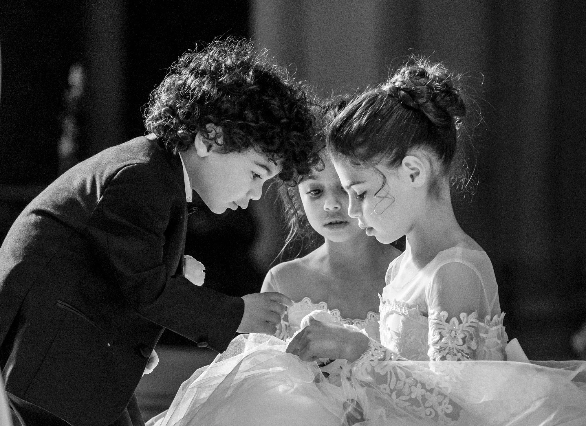 Three children, two girls and one boy, dressed formally, are closely looking at a small item or object, with the girl in a white dress and the boy in a suit, in a black and white photo.