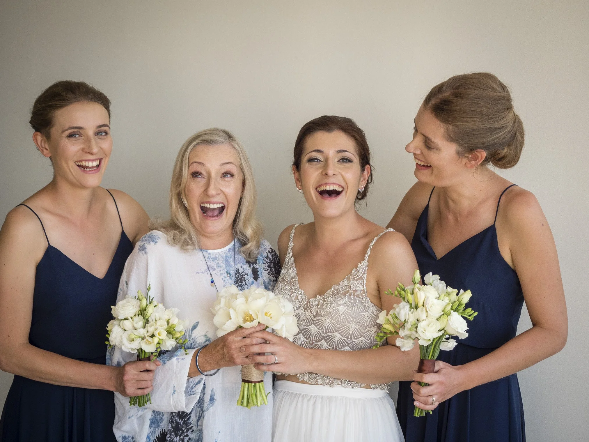 Four women, including a bride in a wedding dress, smiling and holding bouquets, celebrating together.