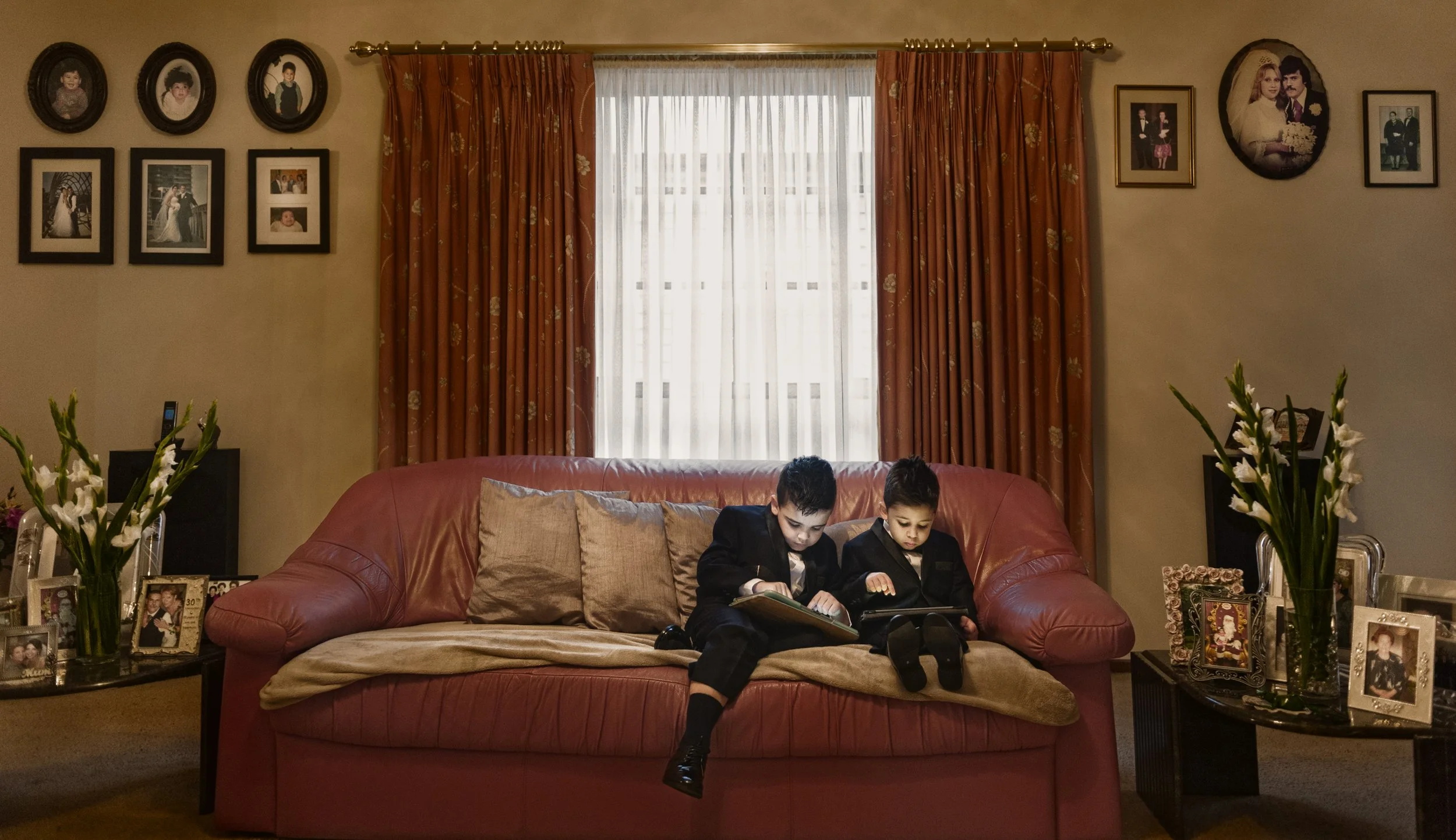 Two young boys dressed in formal black suits sitting on a pink sofa, reading and looking at tablets, in a living room with family photos on side tables and wall.