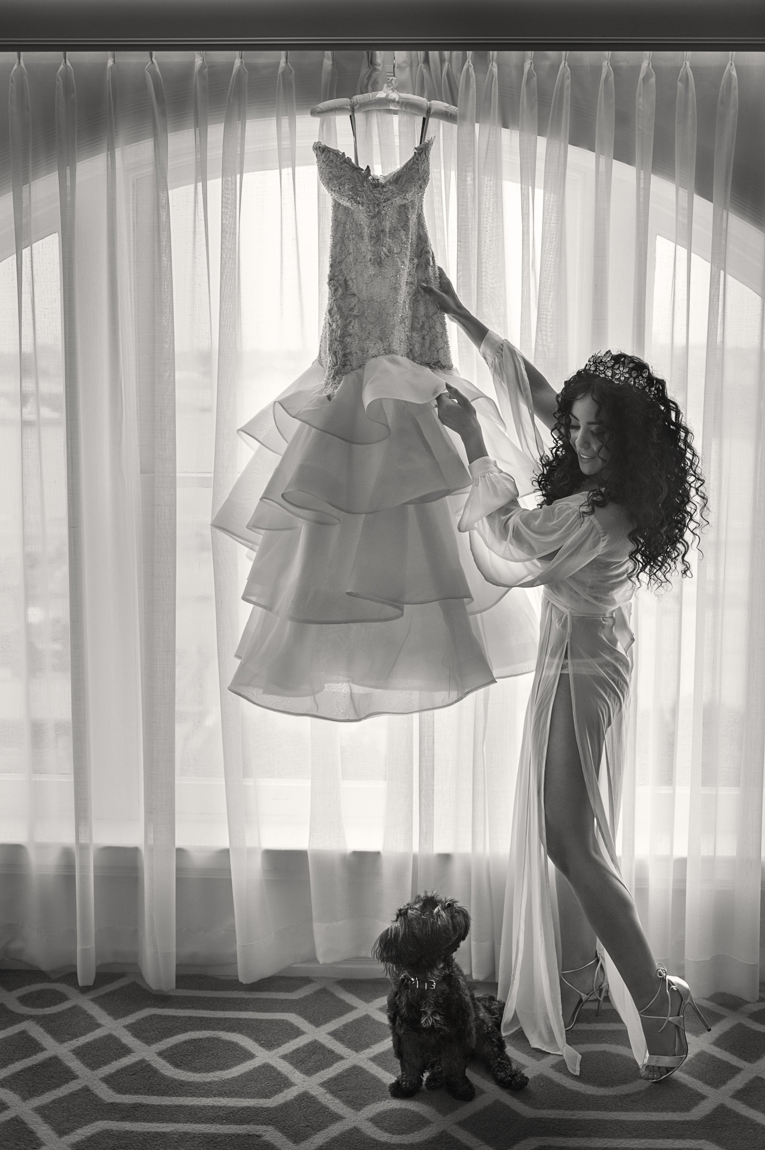 A woman with curly hair and a tiara stands in front of sheer curtains, holding a wedding dress that hangs from a hanger. A small dog sits on the floor looking up at the dress.