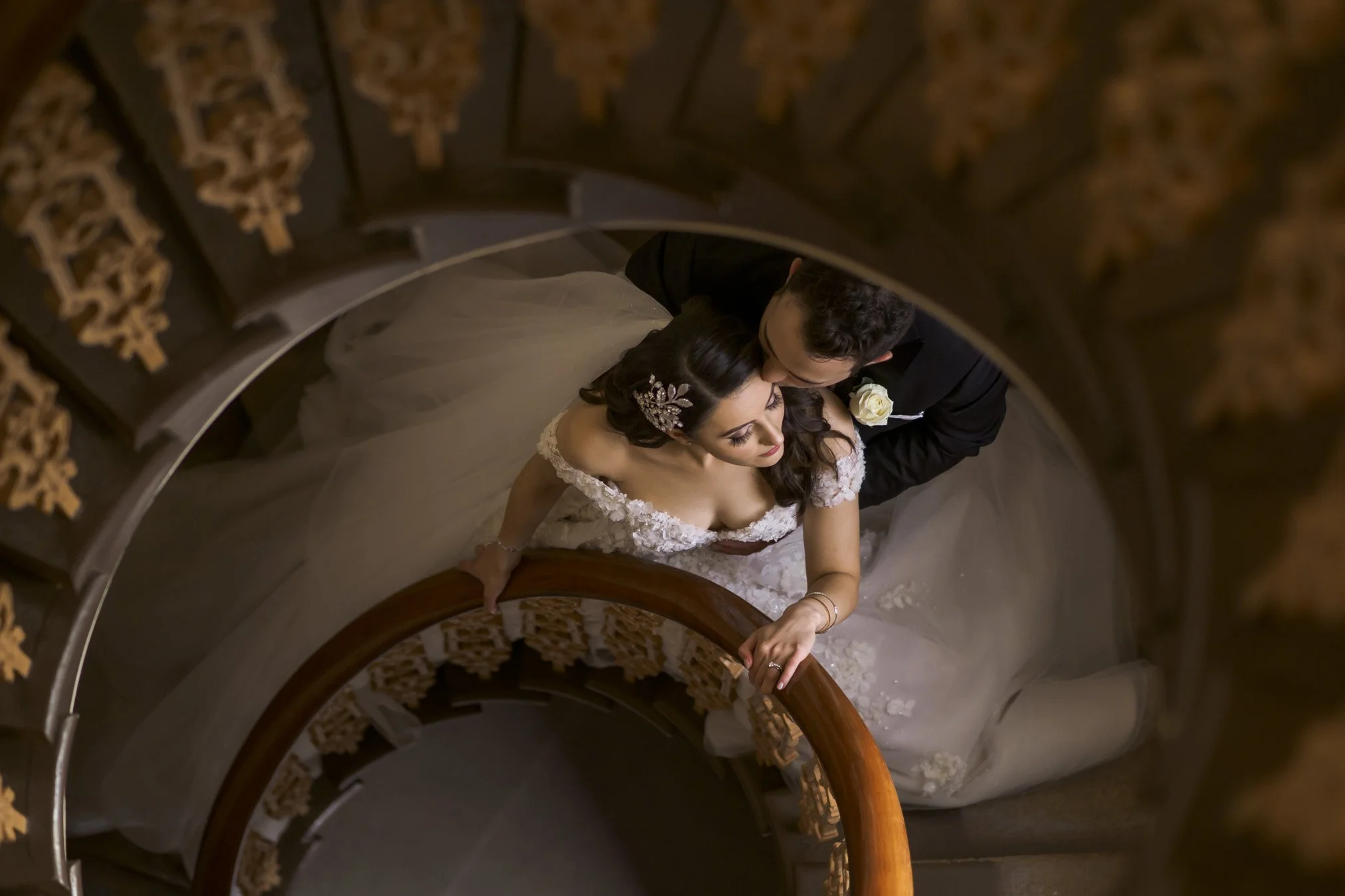 Top-down view of a bride and groom standing on a spiral staircase, with the bride in a white wedding dress and the groom in a black tuxedo, embracing each other.