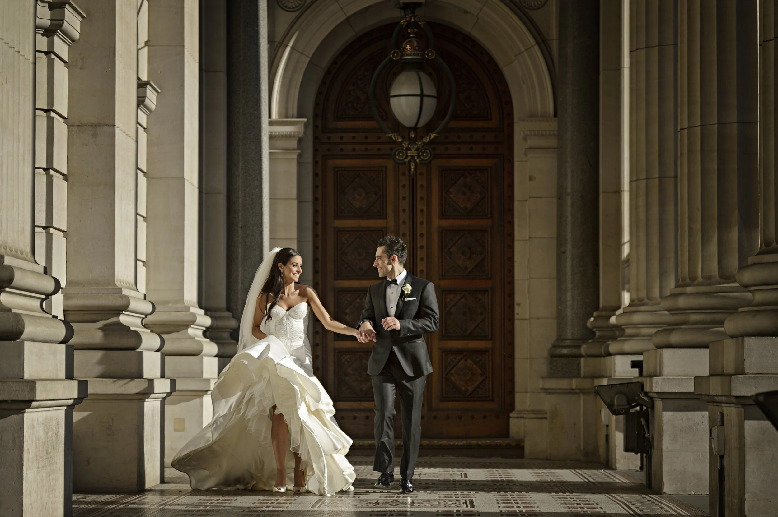 A bride and groom holding hands and walking through a grand, historic building with marble columns and a large wooden door.