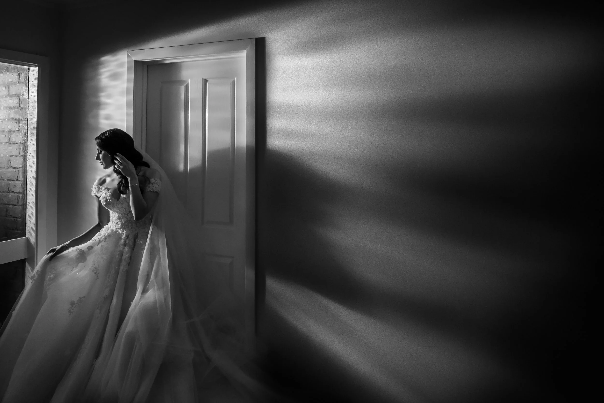 Black and white photo of a bride sitting near a window with light casting shadows on the wall.