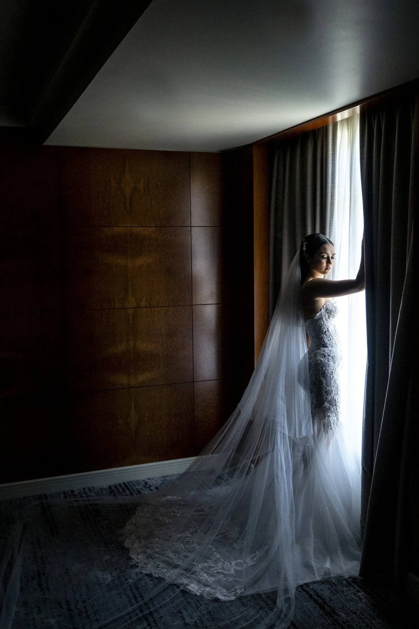 Bride in a lace wedding dress with a long veil standing by a window, holding back curtains as she looks outside in a dimly lit room.