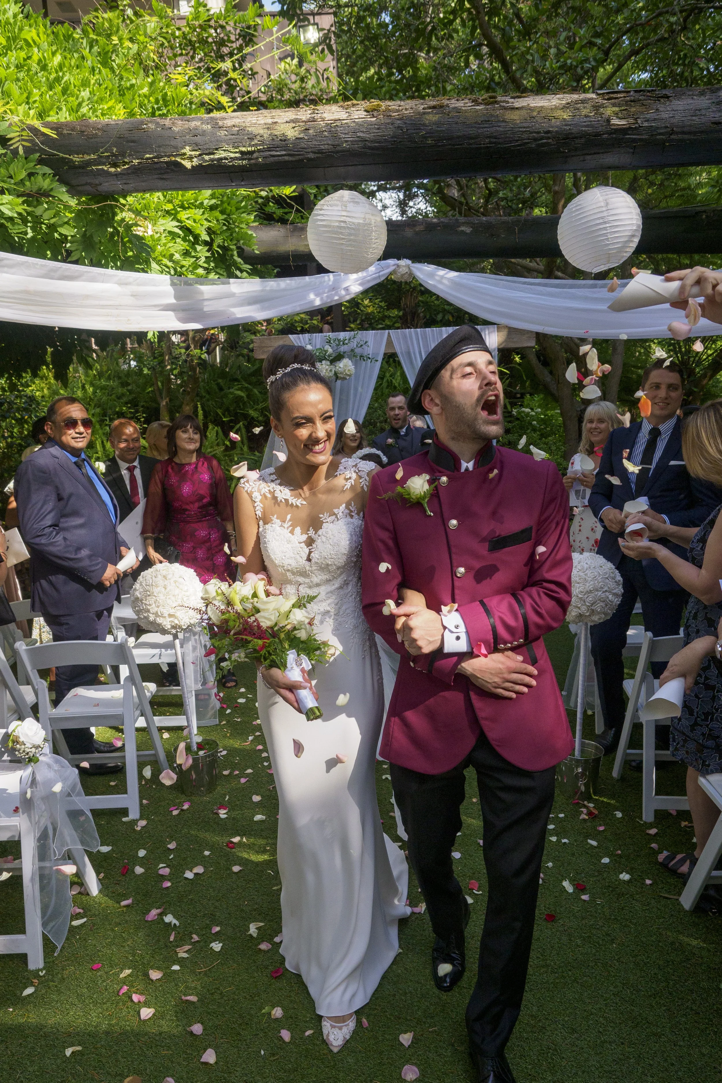 A bride and groom walking down the aisle at their outdoor wedding, surrounded by guests celebrating and throwing flower petals.