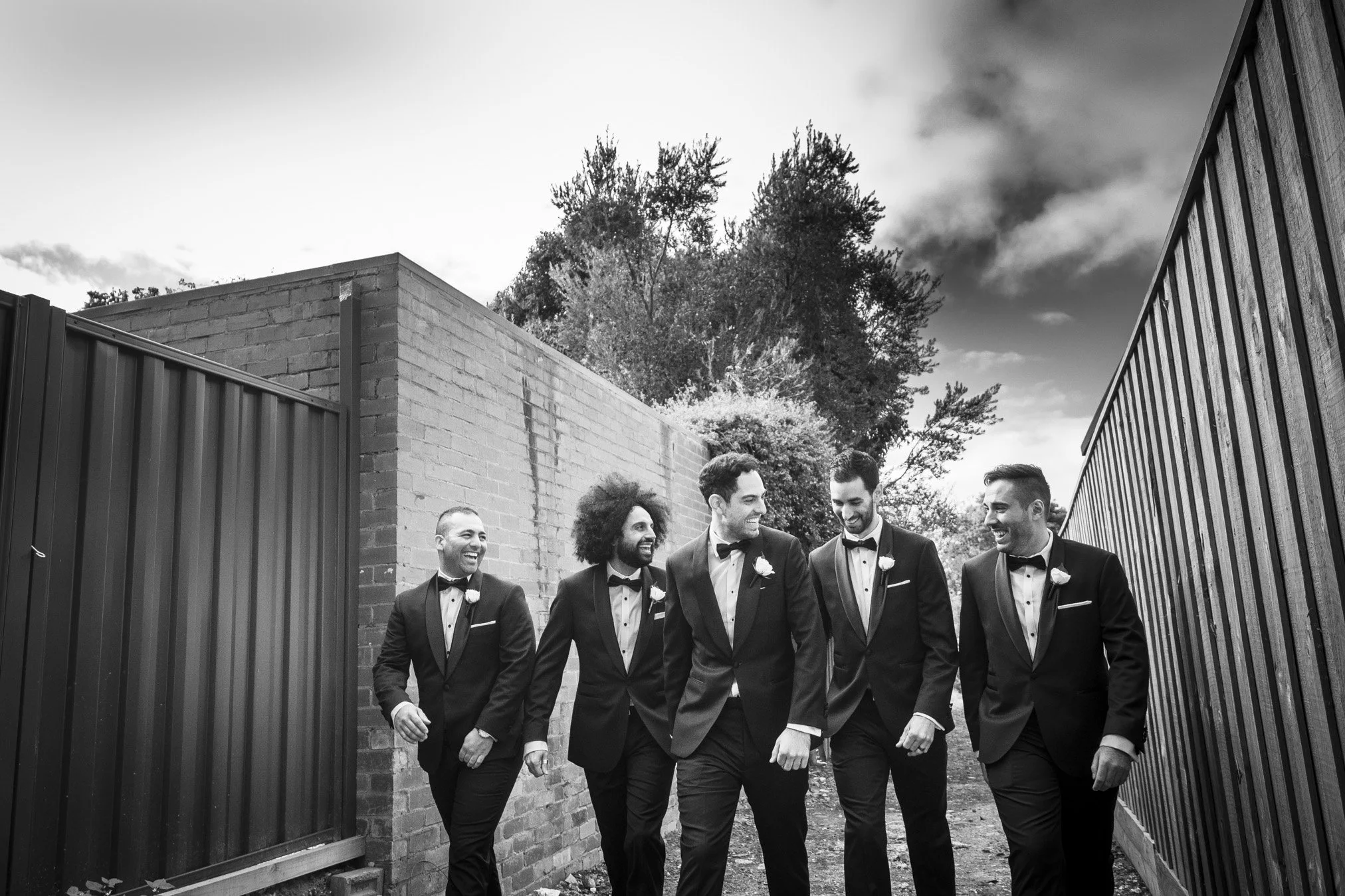A group of five men walking together outdoors, dressed in black tuxedos with white shirts and bow ties, smiling and talking.