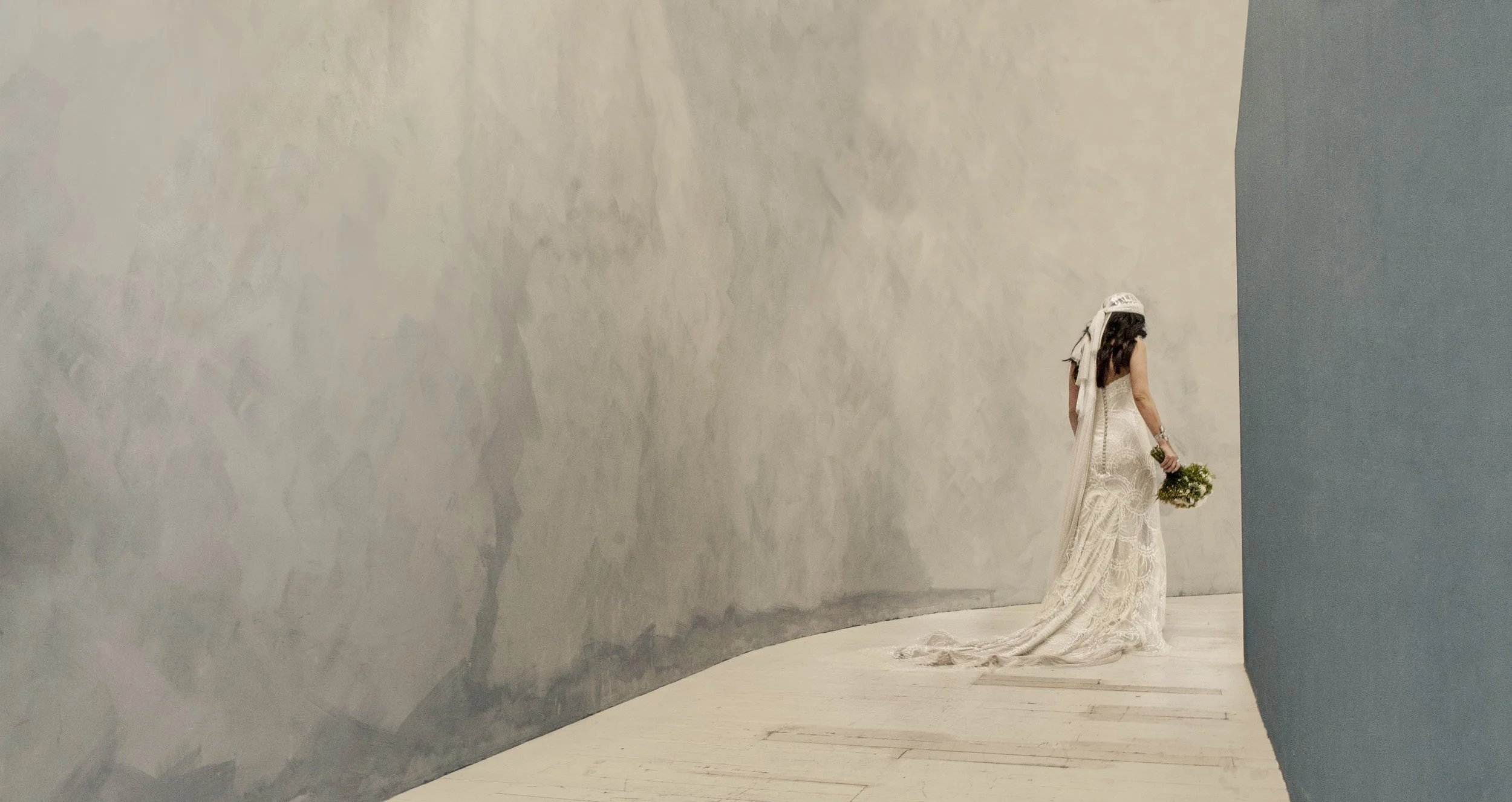 A woman in a wedding dress holding a bouquet walking along a curved white wall, with a blue wall on her right side.