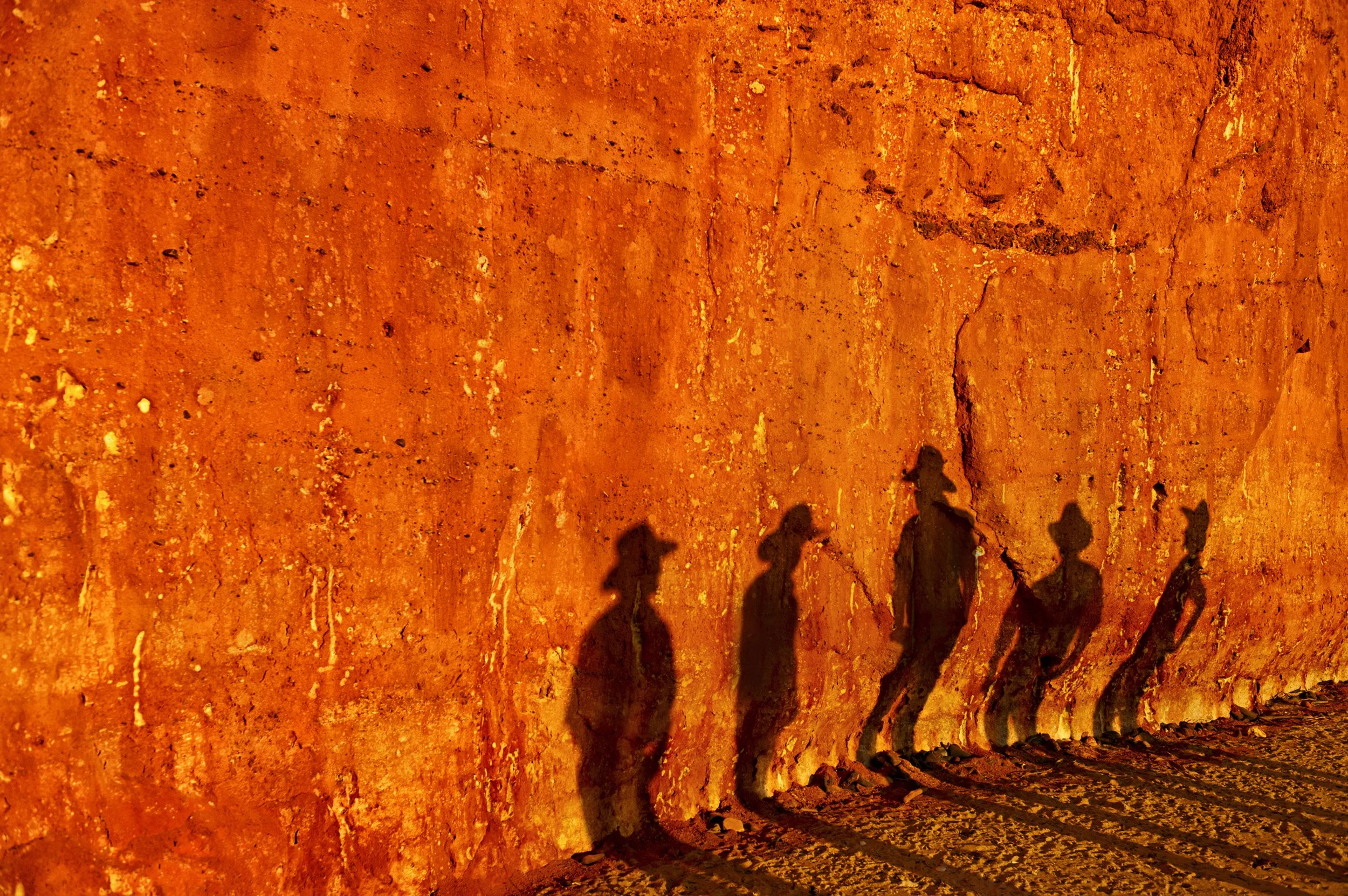 Silhouettes of five people wearing hats and carrying equipment cast shadows on a textured, orange-colored wall.