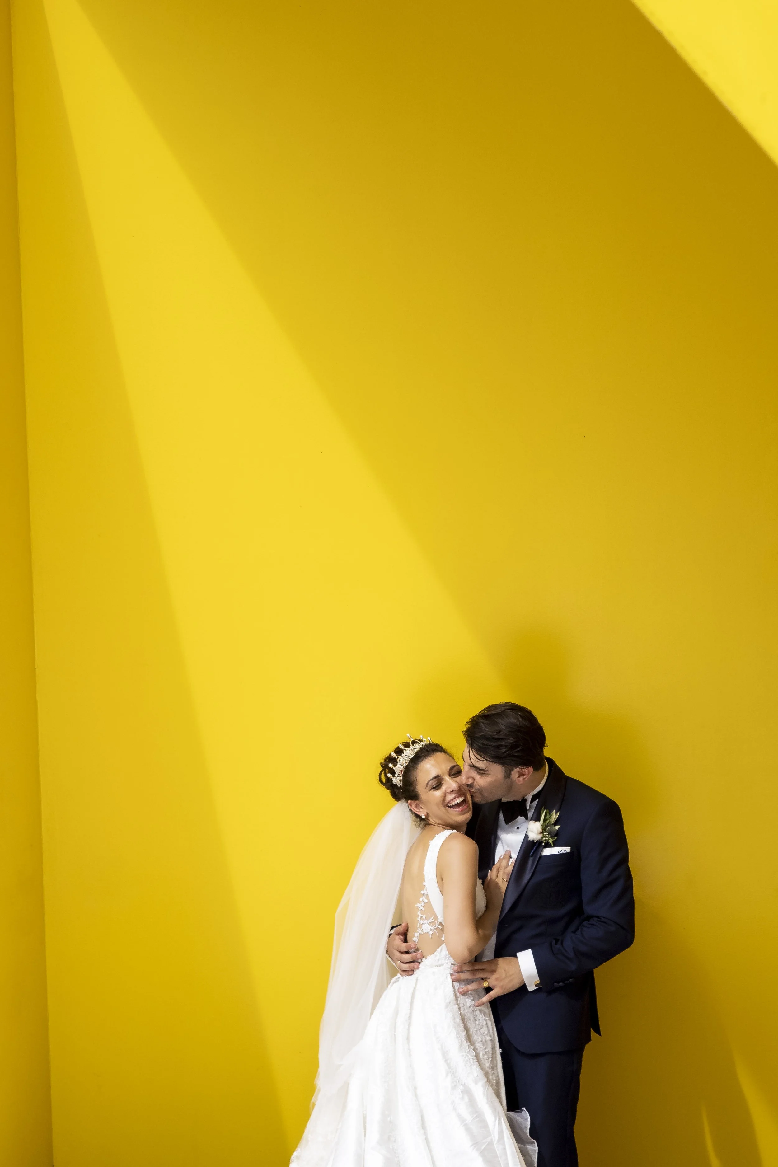 A newlywed couple sharing a joyful moment, with the bride in a white wedding gown and the groom in a navy suit, against a bright yellow background.