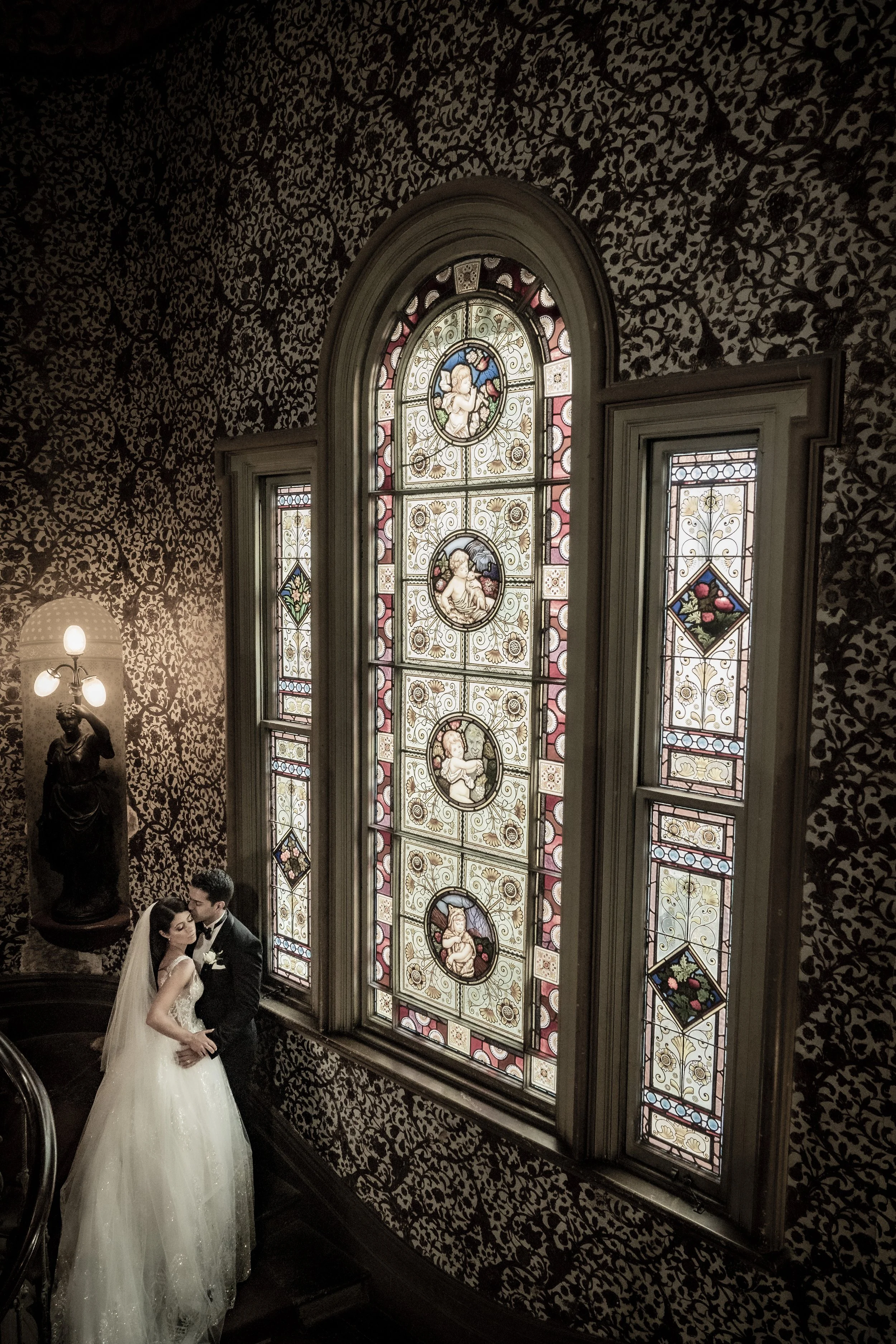 A bride and groom in wedding attire standing close together inside a dimly lit room with intricate, dark patterned wallpaper. Behind them is a large, colorful stained glass window with floral and angelic designs. A statue of a woman holding a lamp is