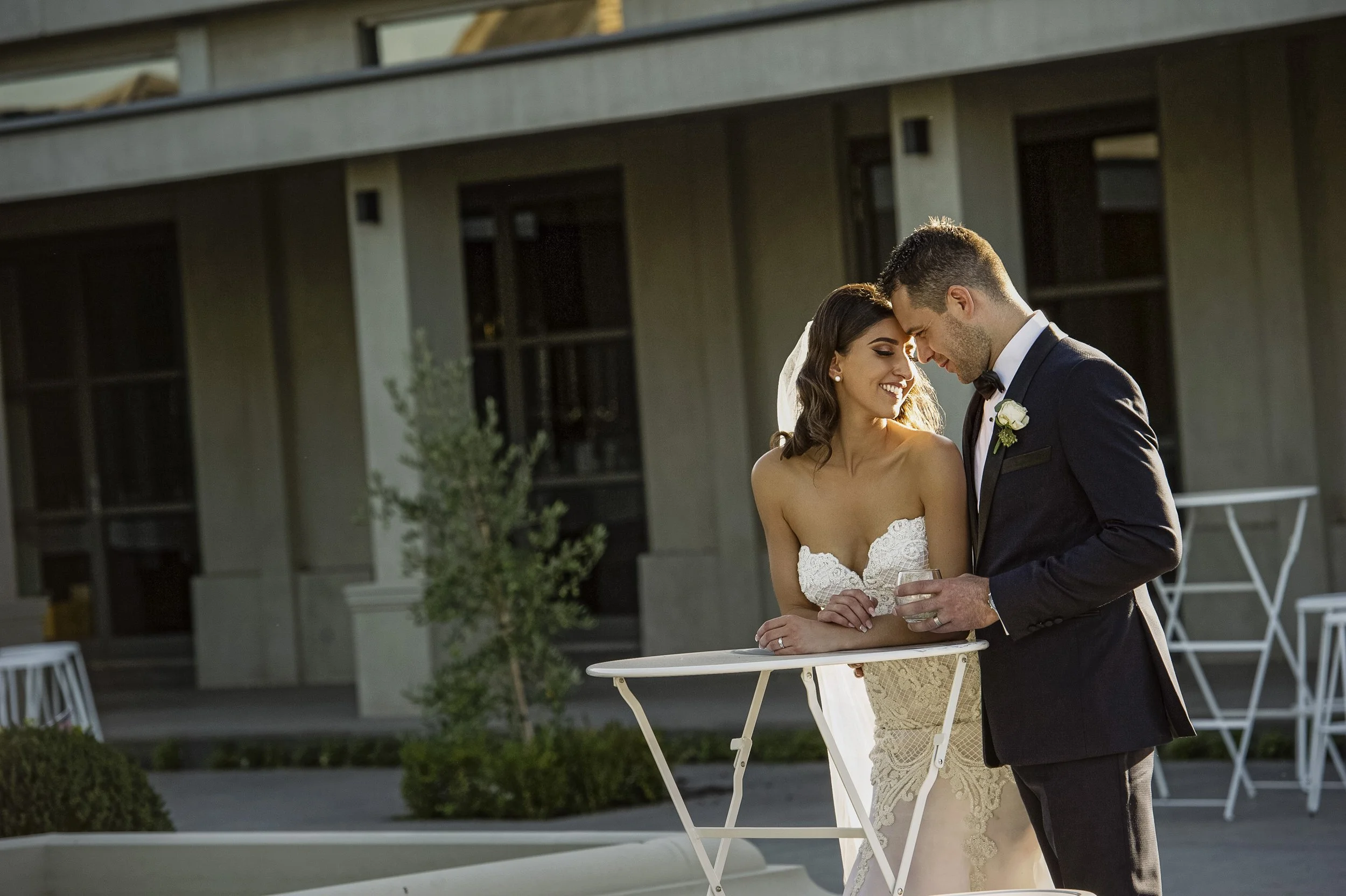 A newlywed couple stands close together outside a building, smiling and gazing at each other. The bride wears a strapless wedding dress with lace details and the groom is in a black tuxedo with a boutonnière. They are leaning on a small white table w