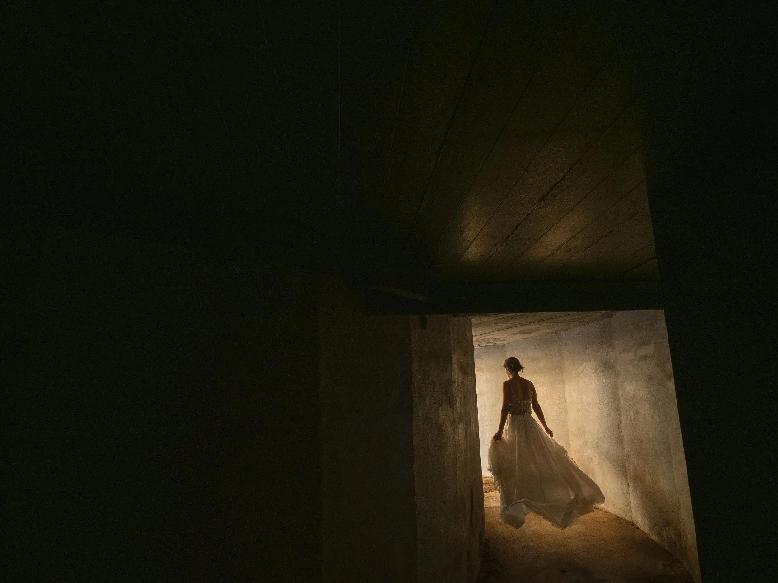 A woman in a wedding dress walking into a dimly lit, concrete room.