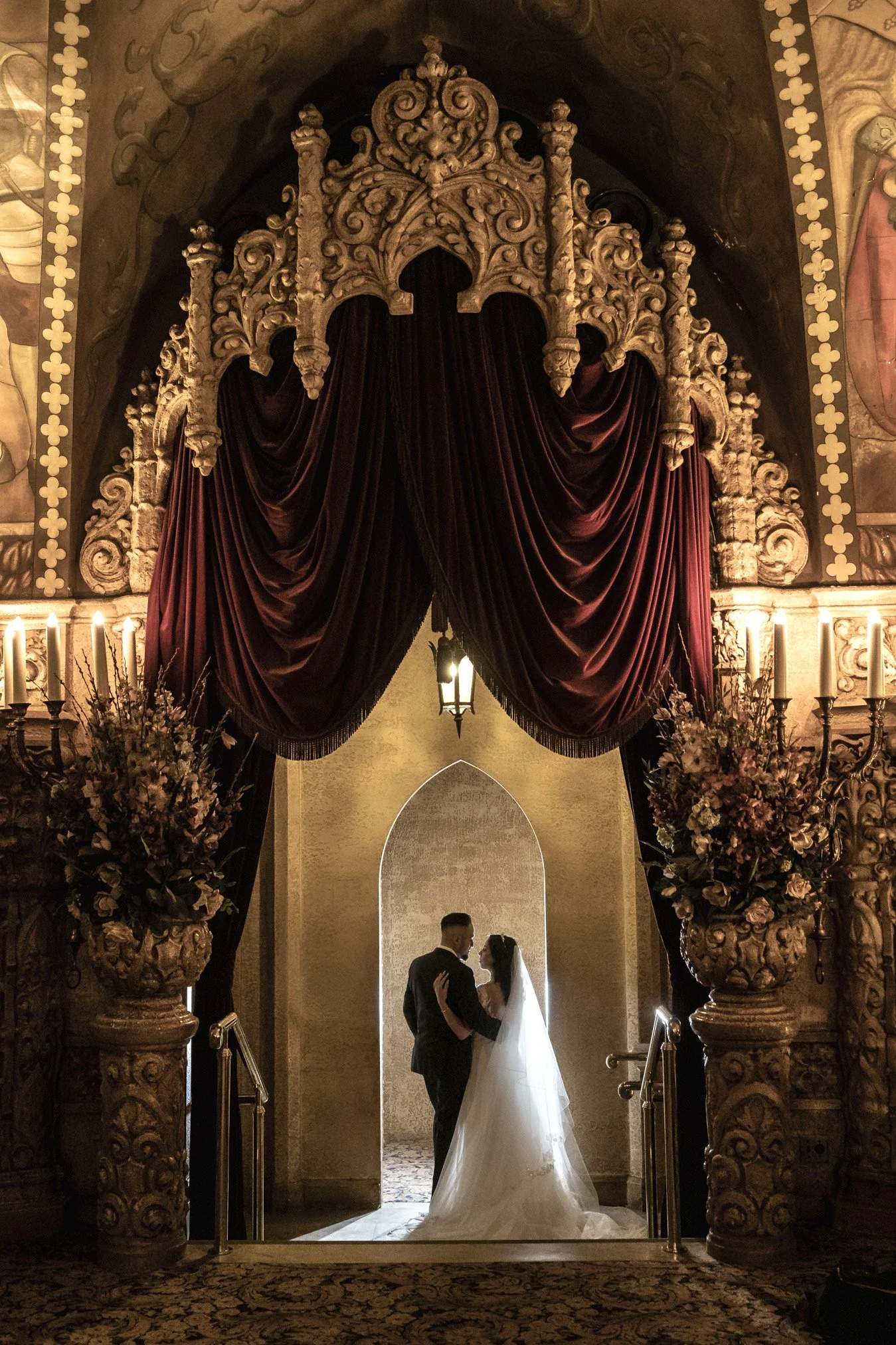 A bride and groom standing close together in a dimly lit, ornate wedding venue, framed by a large archway with draped dark red velvet curtains, floral arrangements, and candles.