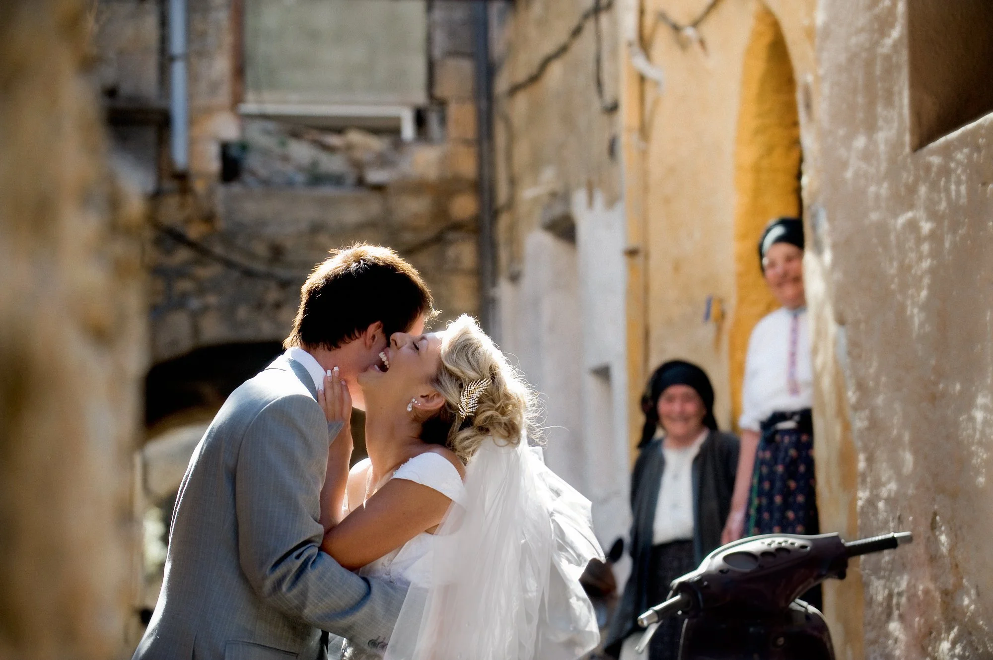 A couple in wedding attire sharing a kiss in a narrow, rustic alleyway, with two women watching and smiling in the background.