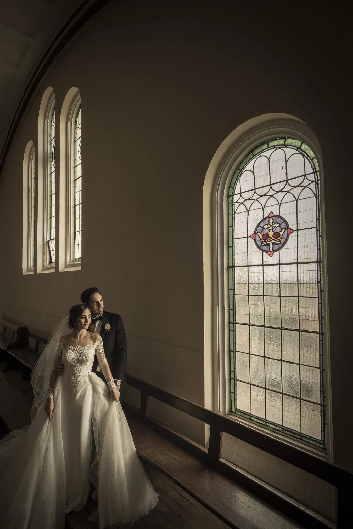 Bride and groom standing inside a church near tall stained glass windows, with the bride wearing a white wedding gown and veil, and the groom in a black tuxedo.