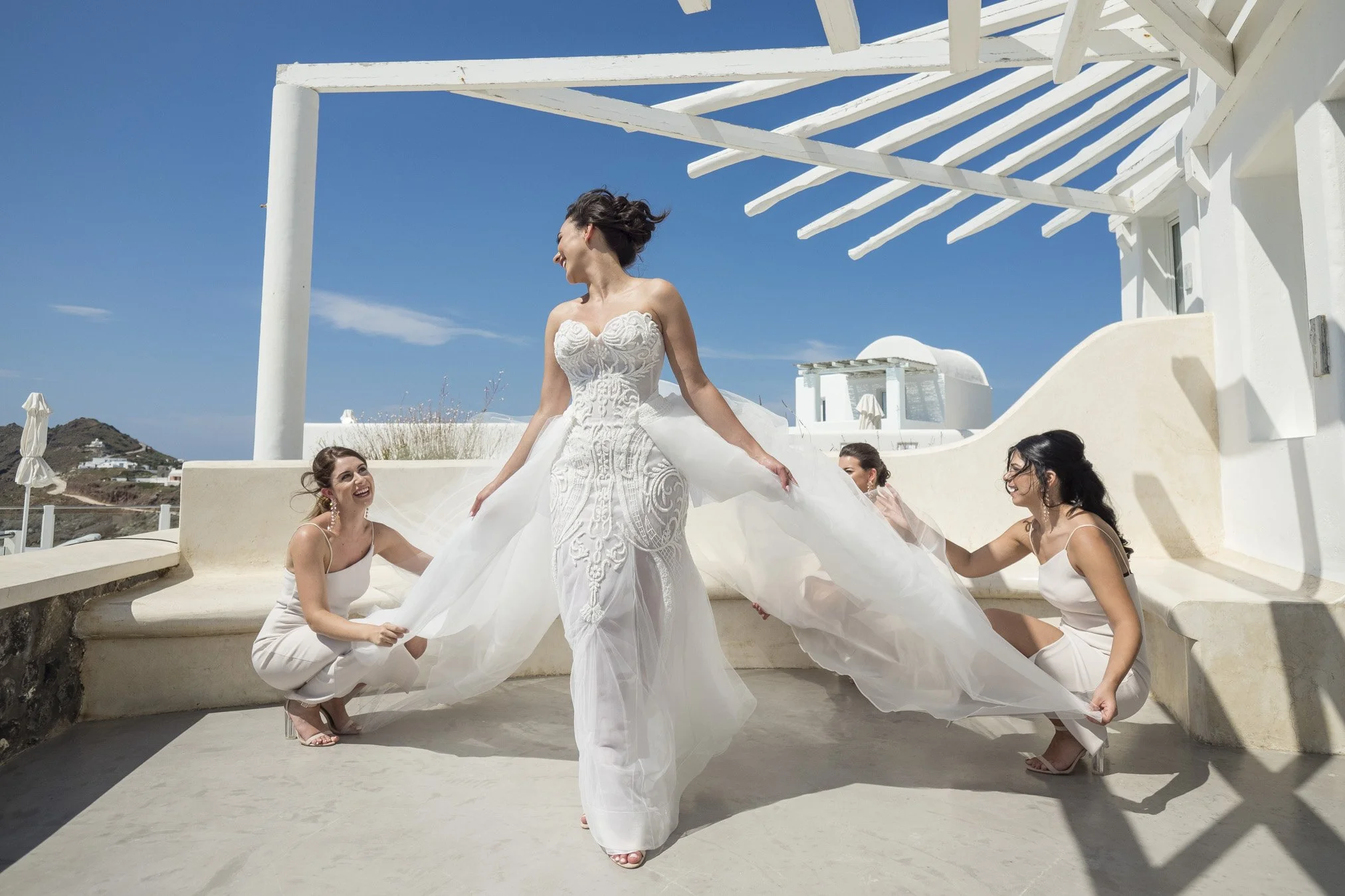 A bride in a white wedding gown stands holding the sides of her dress while four bridesmaids in white dresses help her with her dress outside under a white pergola on a sunny day, with white buildings and a hill in the background.