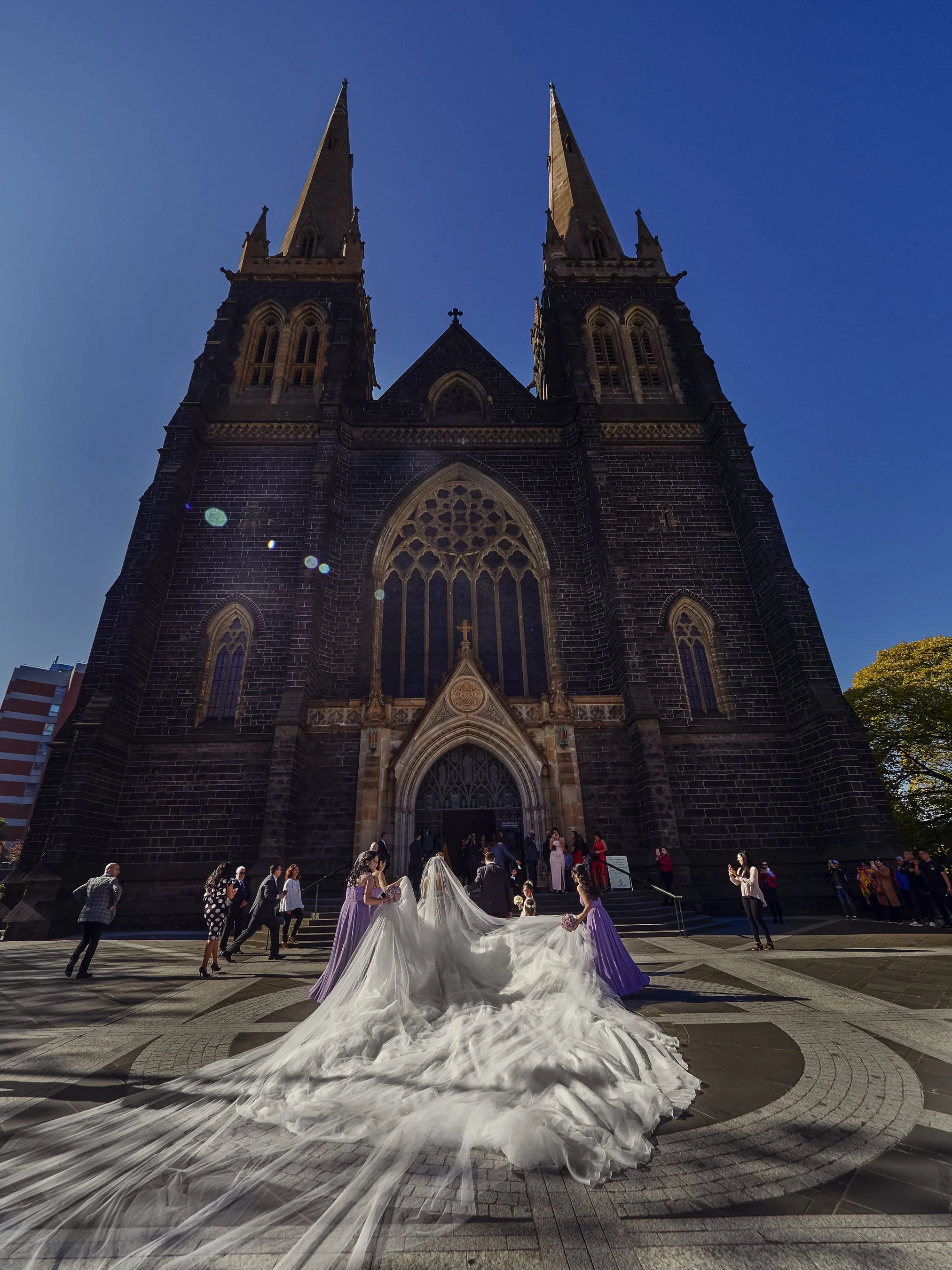 A bride in a white wedding gown with a long train, standing in front of a large Gothic-style church with twin spires on a sunny day. She is surrounded by bridesmaids and wedding guests.