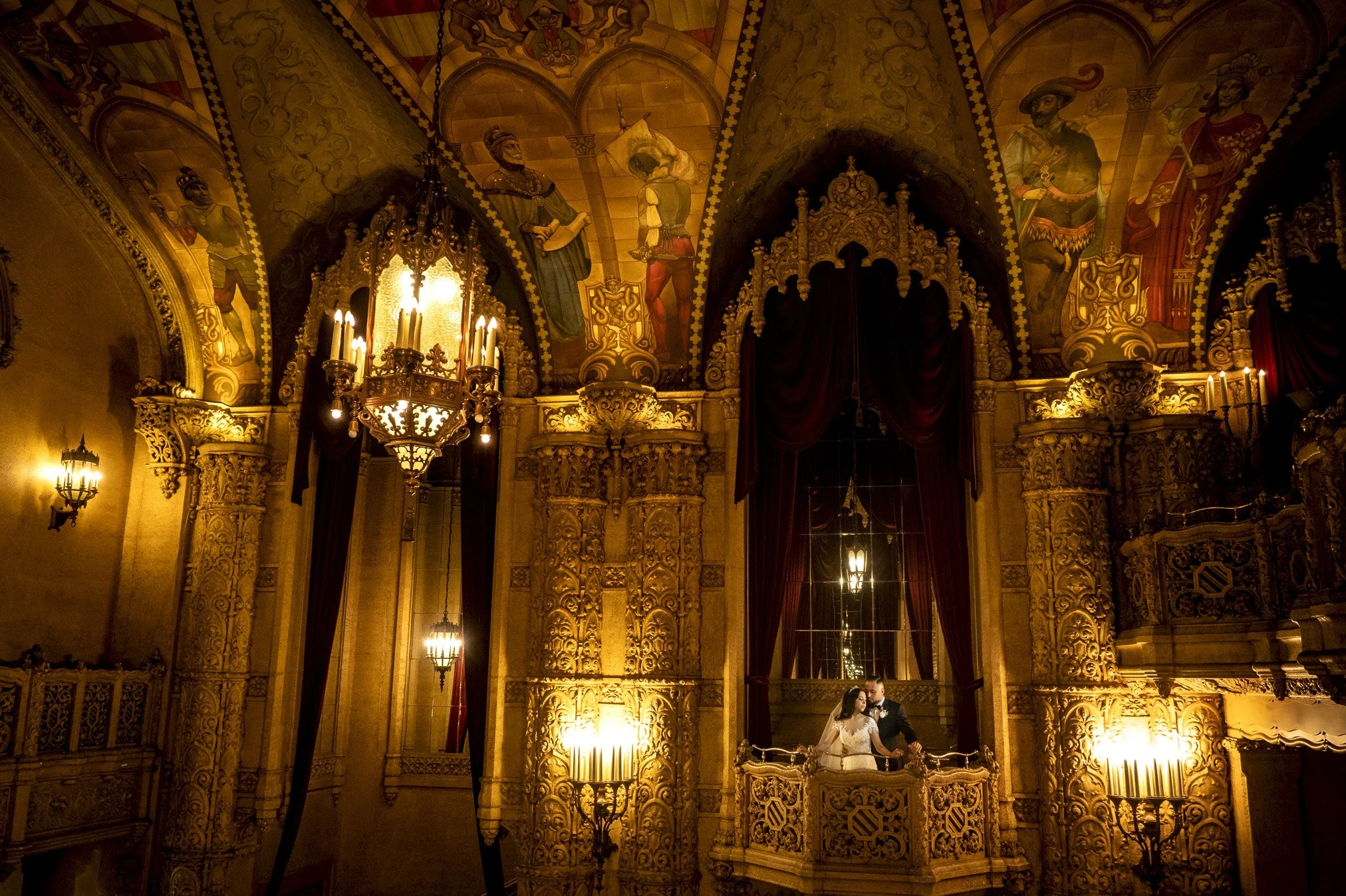 A couple in wedding attire standing on a grand, ornate balcony inside a richly decorated, gold-accented hall with chandeliers, frescoes, and dark red curtains.