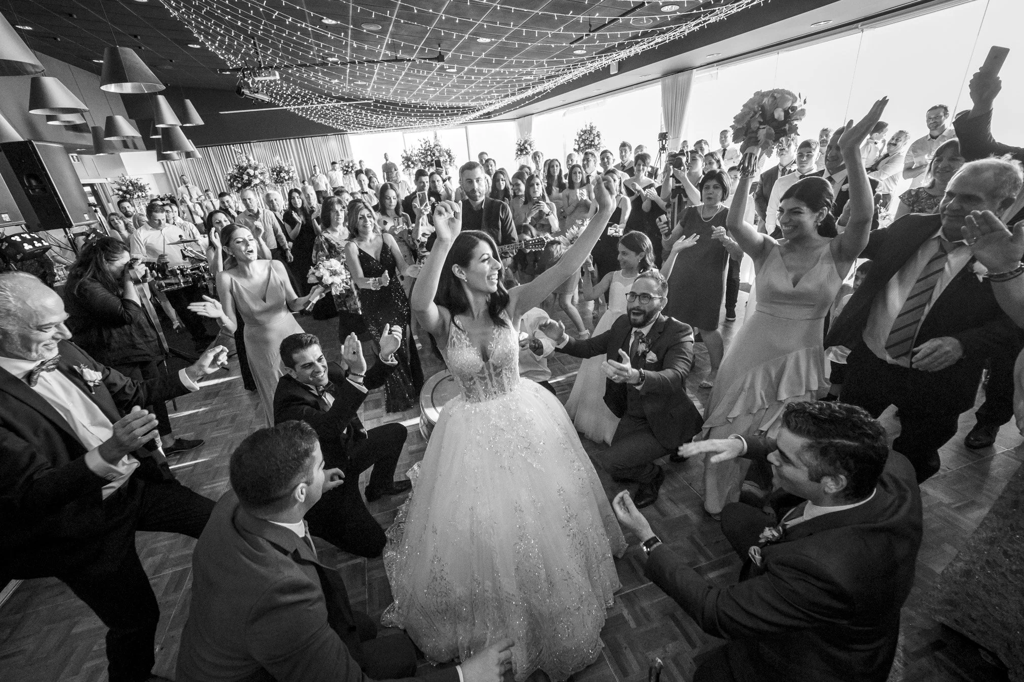 A bride and groom dancing at their wedding reception surrounded by guests, with many people smiling, clapping, and taking pictures in a decorated banquet hall.