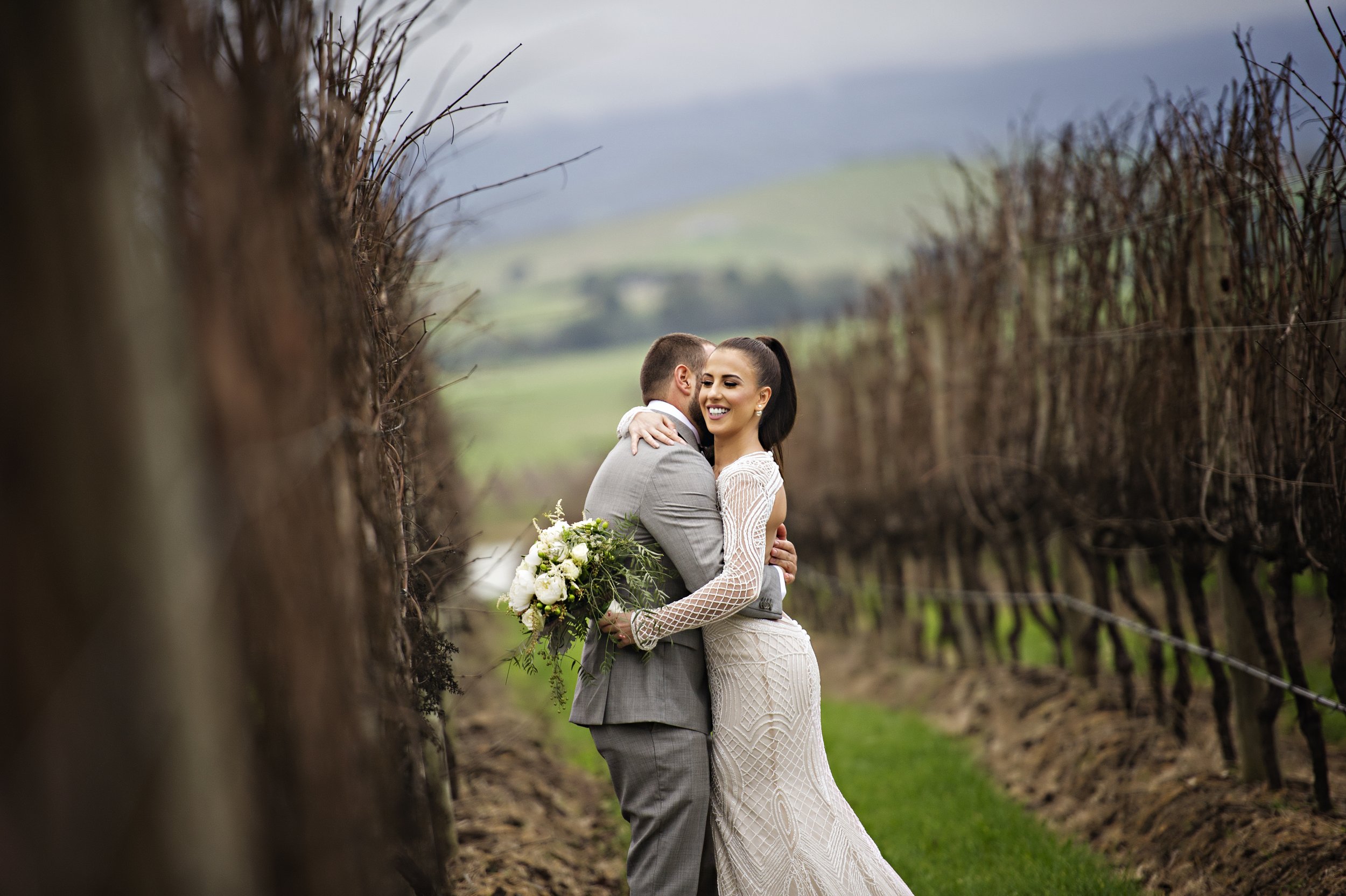 A bride and groom embrace in a vineyard, with the bride holding a bouquet of white flowers, as green hills and a cloudy sky are visible in the background.