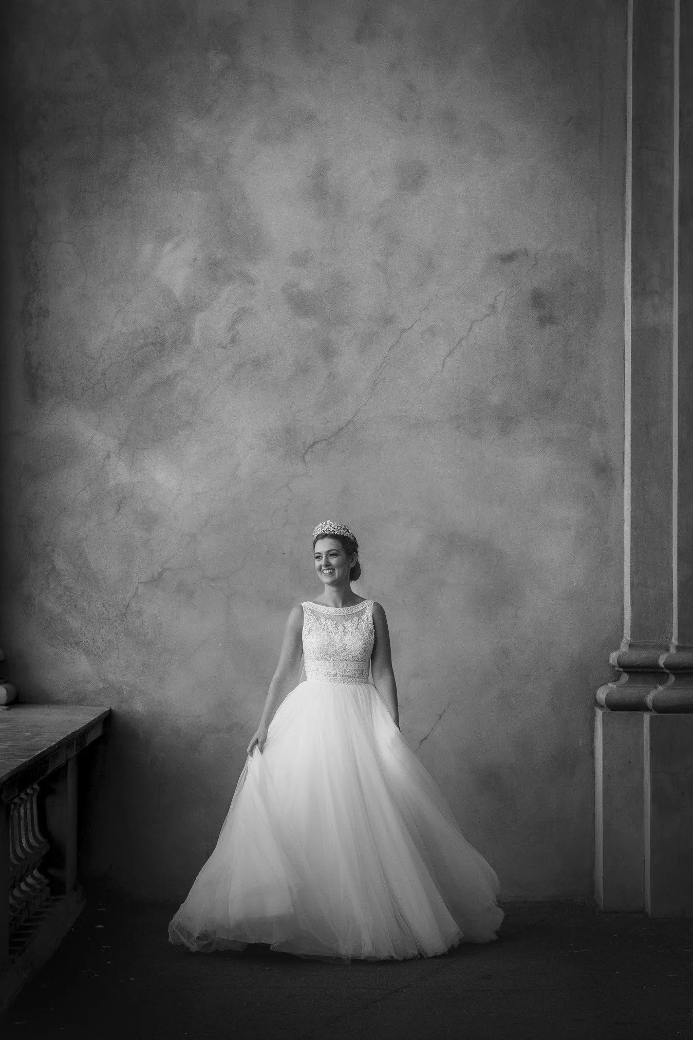 A woman in a wedding dress and tiara smiling, standing against a textured wall.