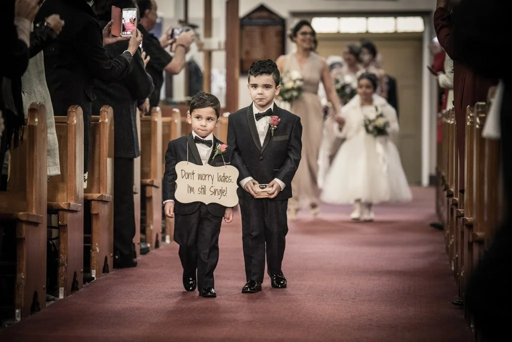 Two young boys dressed in tuxedos walking down an aisle in a wedding ceremony, with people taking photos and a bride walking behind them.