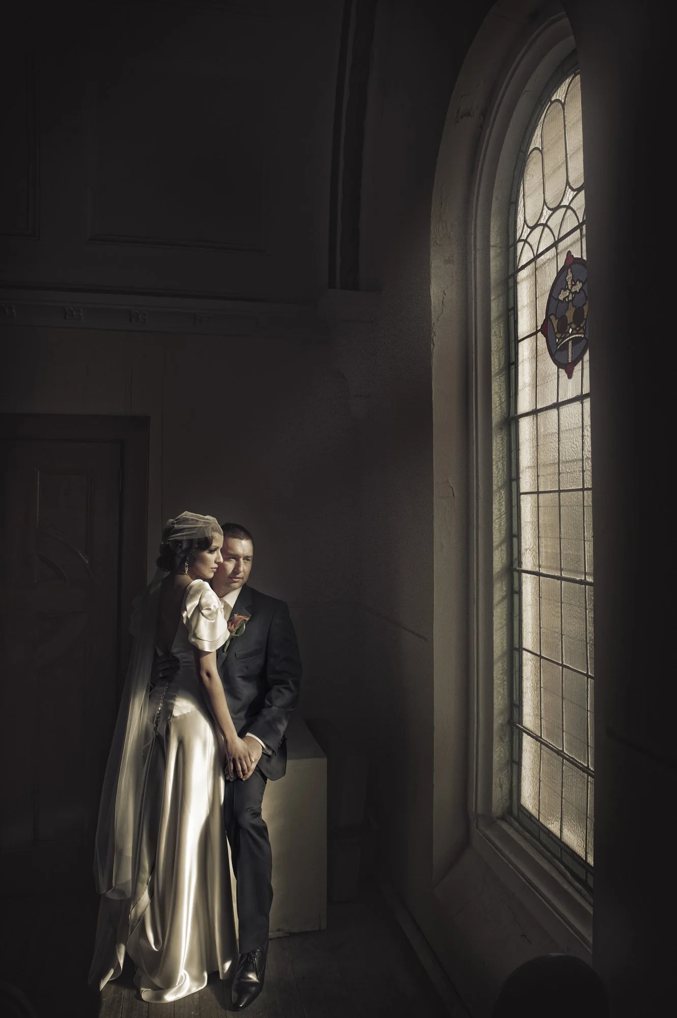 Bride and groom sitting together inside a dimly lit room near a large stained glass window.