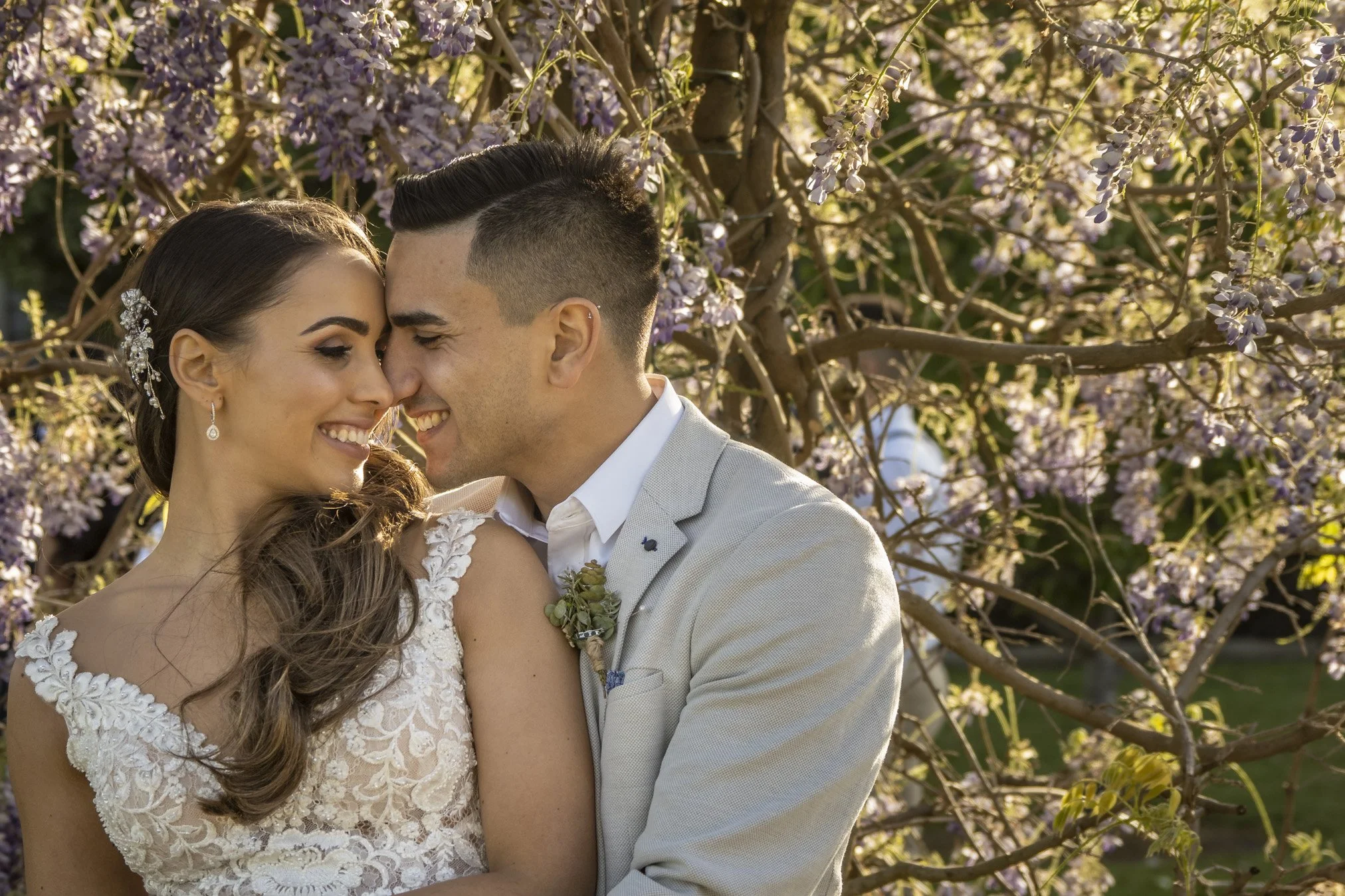 A happy couple on their wedding day, standing close with their foreheads touching, surrounded by blooming purple flowers.