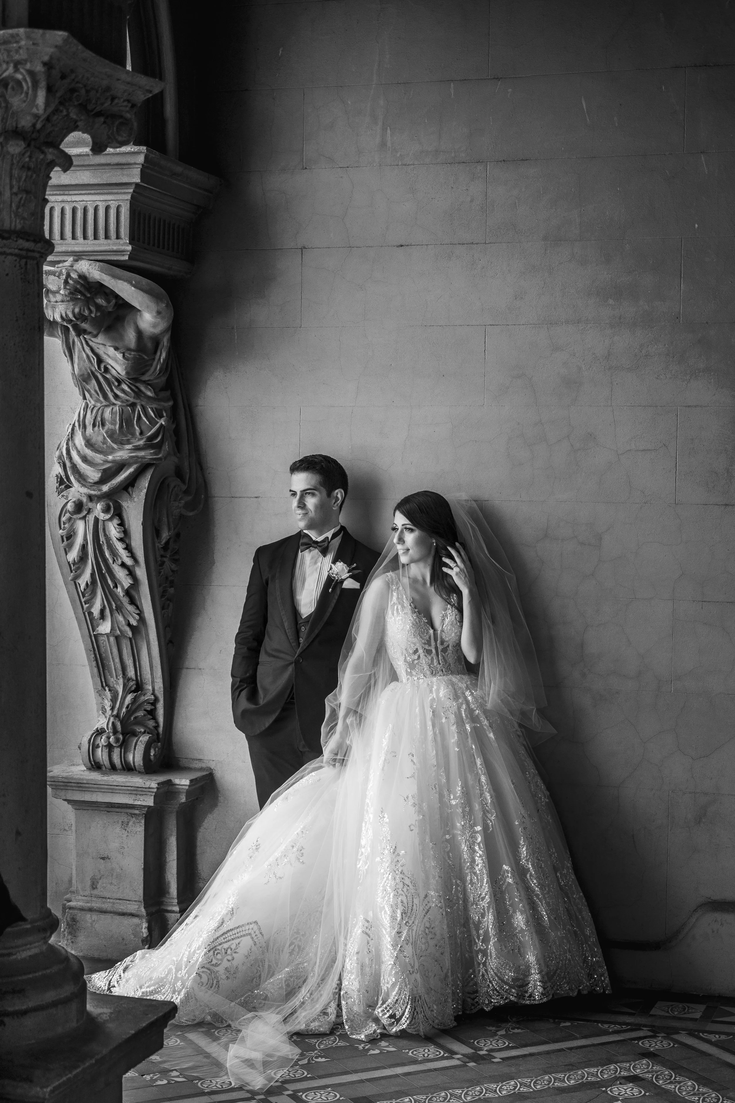 Black and white photograph of a bride in an ornate wedding gown and veil standing next to a groom in a tuxedo, both leaning against a decorated wall inside a grand building.