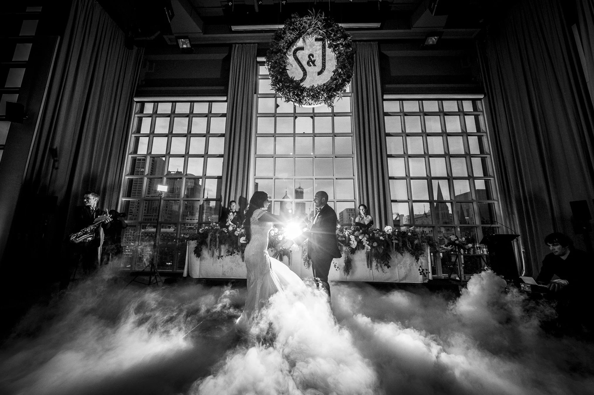 A black-and-white photo of a wedding ceremony indoors featuring a couple dancing in front of a large window with a city skyline view. The bride and groom are holding hands, and there are two women behind them, likely the bridal party, standing at a d