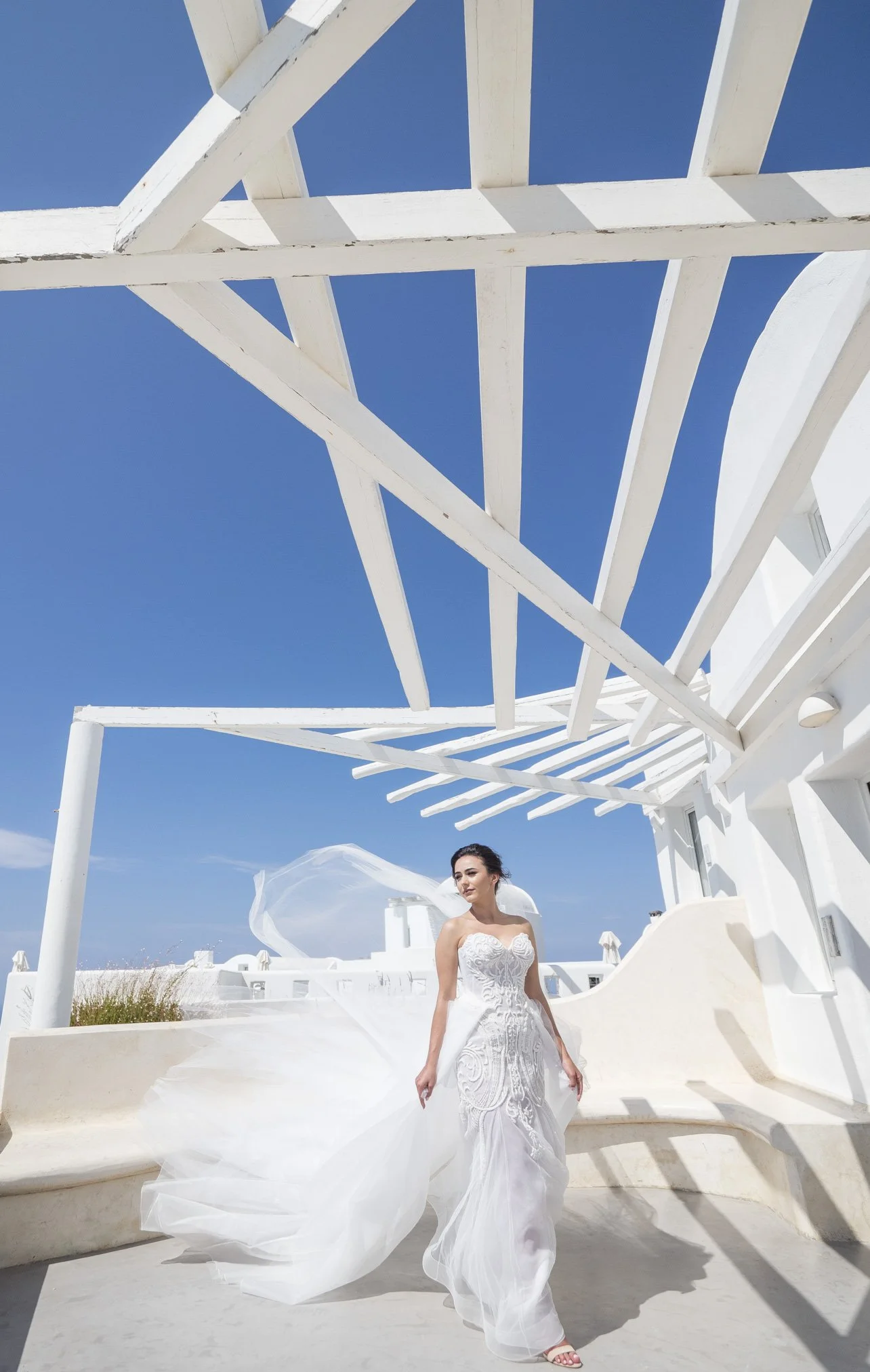 A woman in a white wedding dress standing under a white wooden pergola on a sunny day with a clear blue sky.