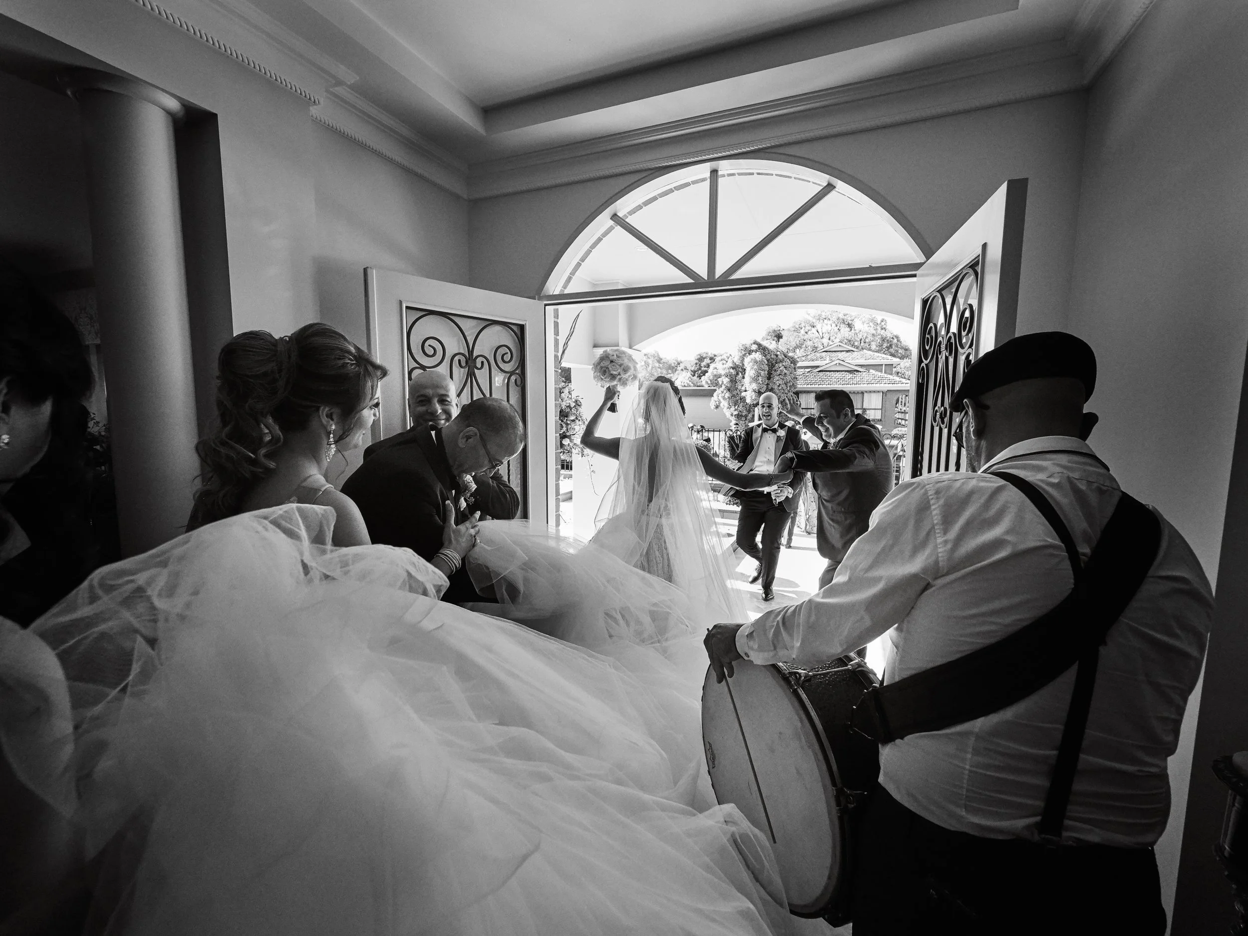 Black-and-white photo of a bride and wedding guests dancing at the doorway of a house, with a musician playing a drum in the foreground.