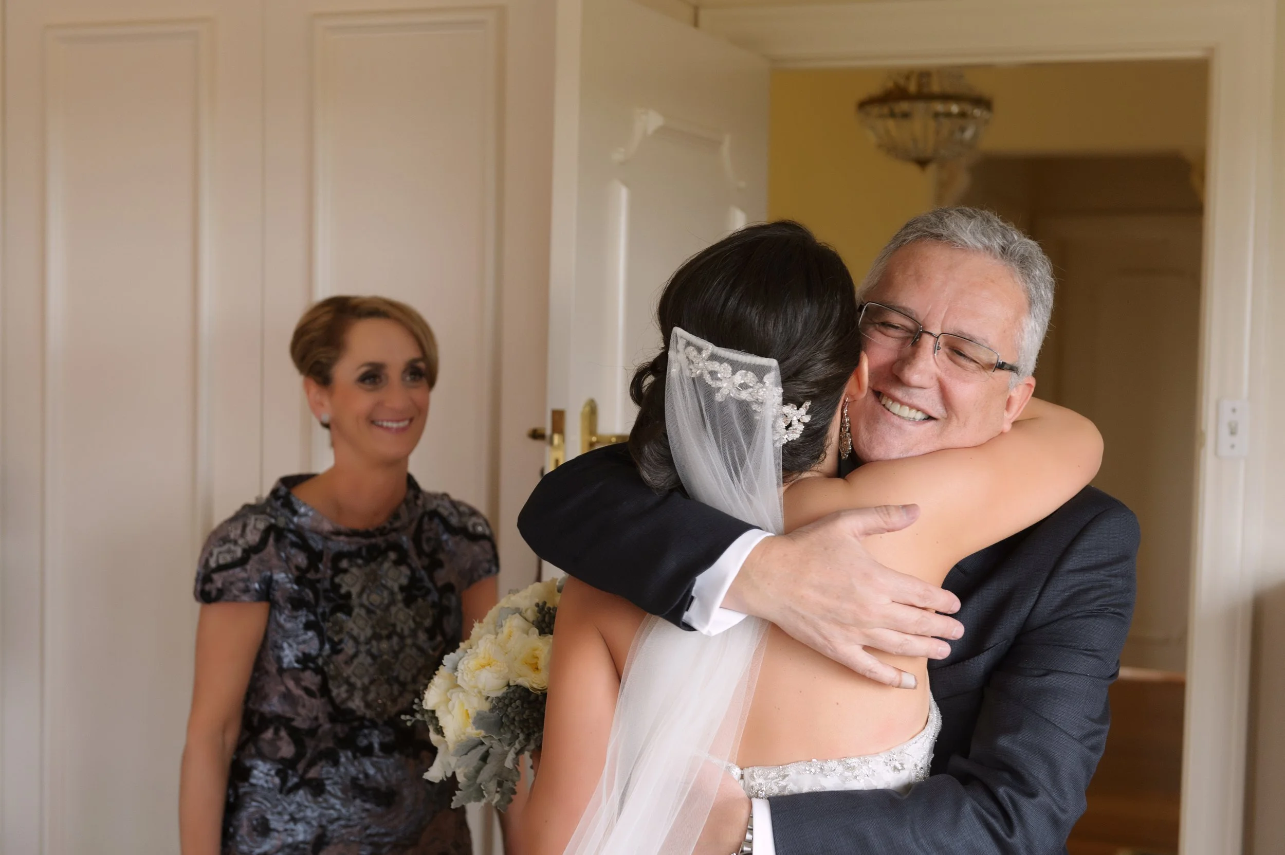 Bride hugging an older man, possibly her father, while a woman watches and smiles in the background.