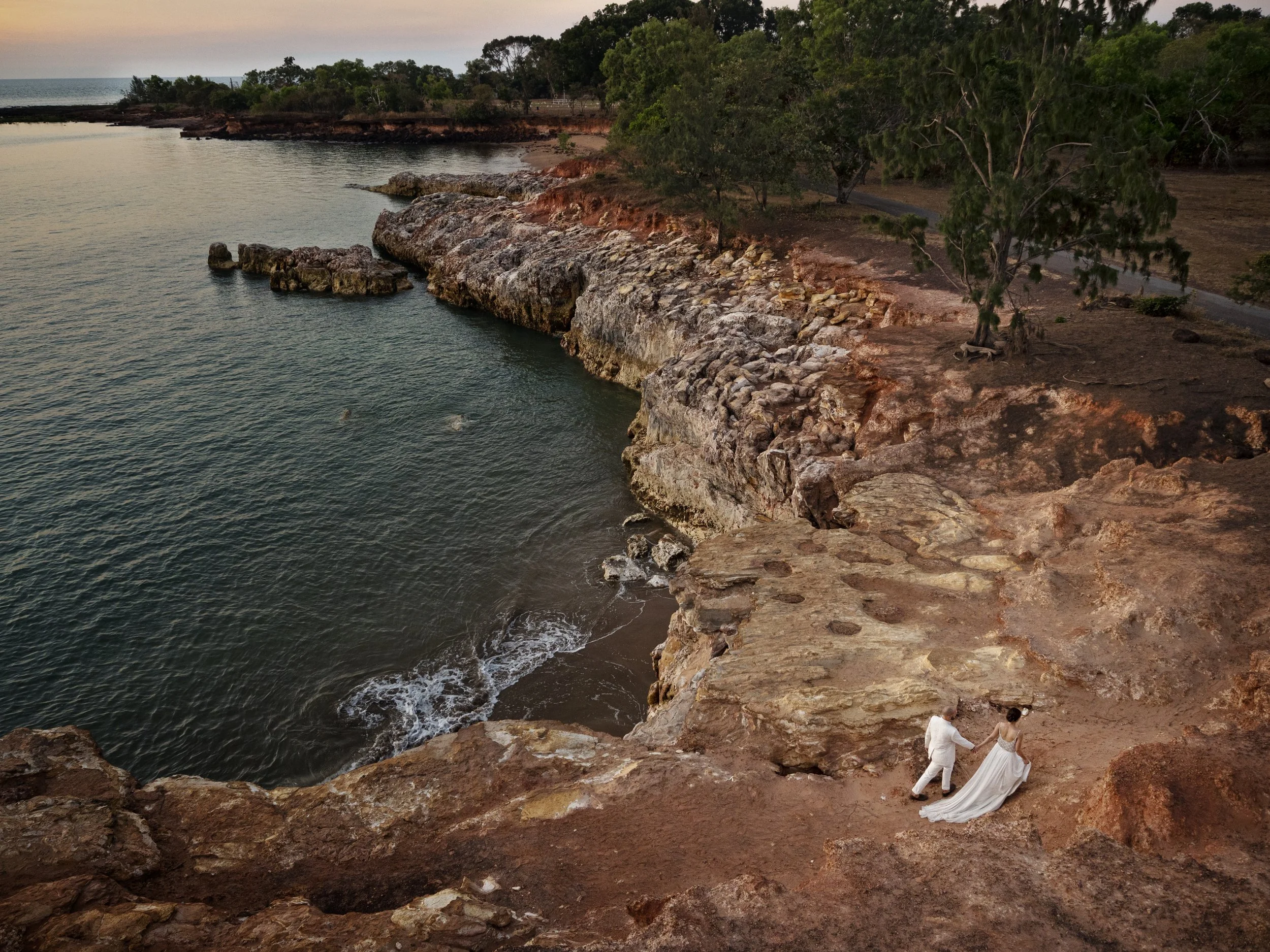 A couple dressed in wedding attire walking on a rocky cliffside overlooking the ocean at sunset.