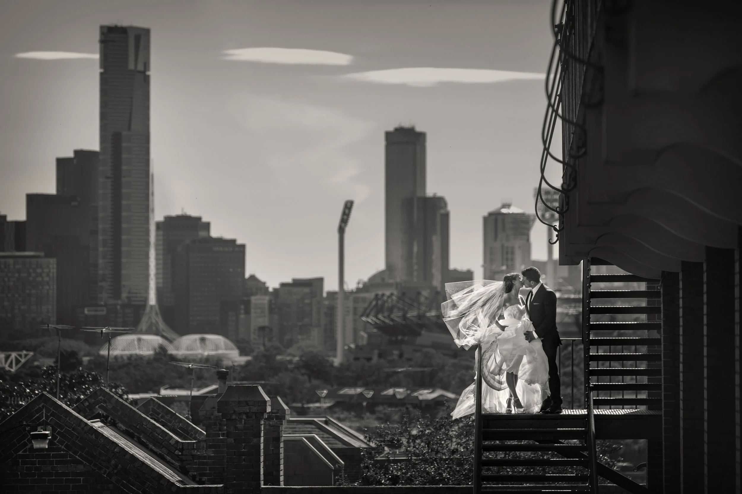 A black and white photo of a bride and groom sharing a kiss on a balcony with an urban cityscape in the background, including tall buildings and skyscrapers.